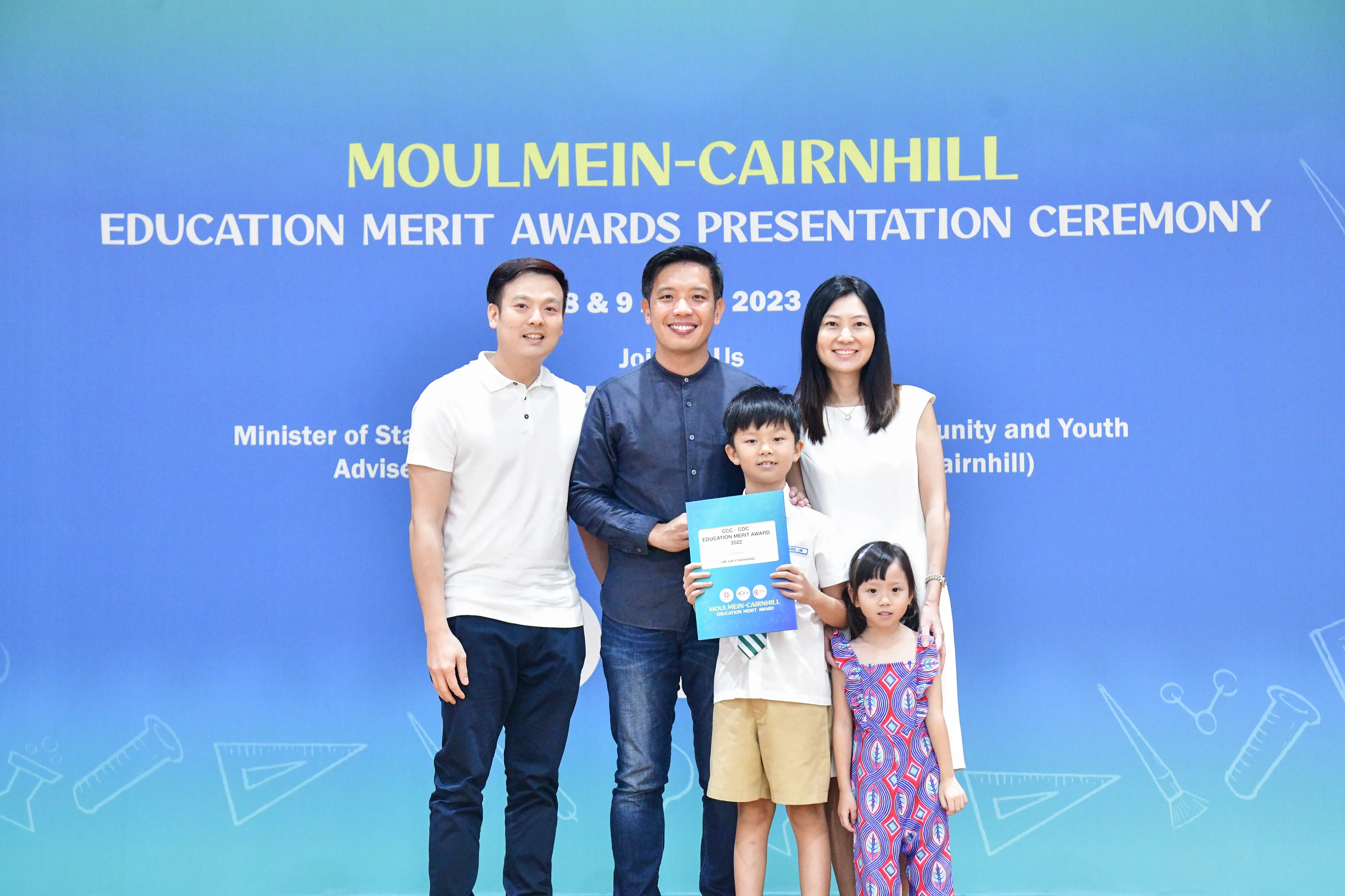 Five people pose before a Moulmein-Cairnhill Education Merit Awards 2023 banner, a boy holding a blue certificate.