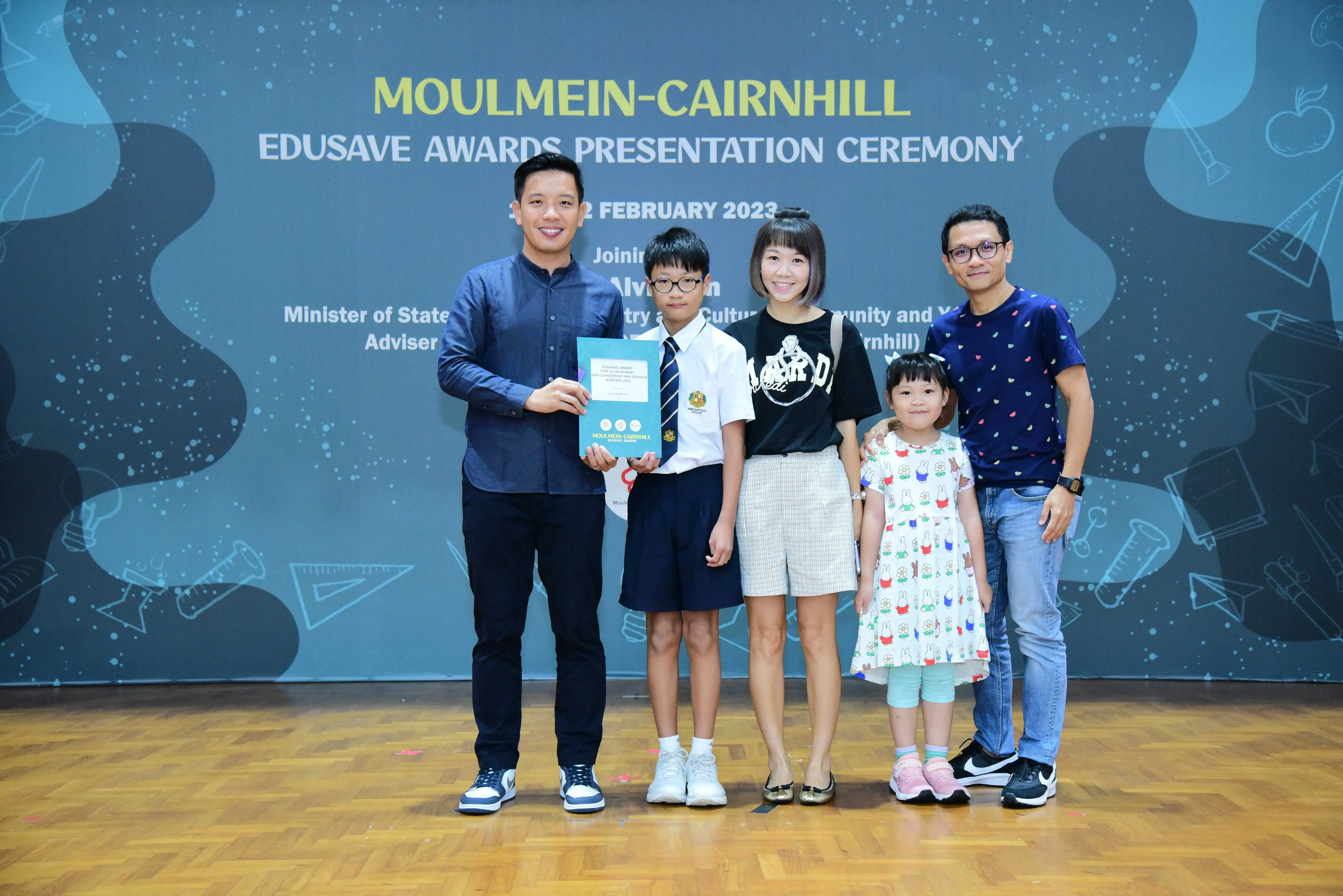 Five people pose in front of a Moulmein-Cairnhill Edusave Awards Presentation Ceremony banner, holding a certificate.