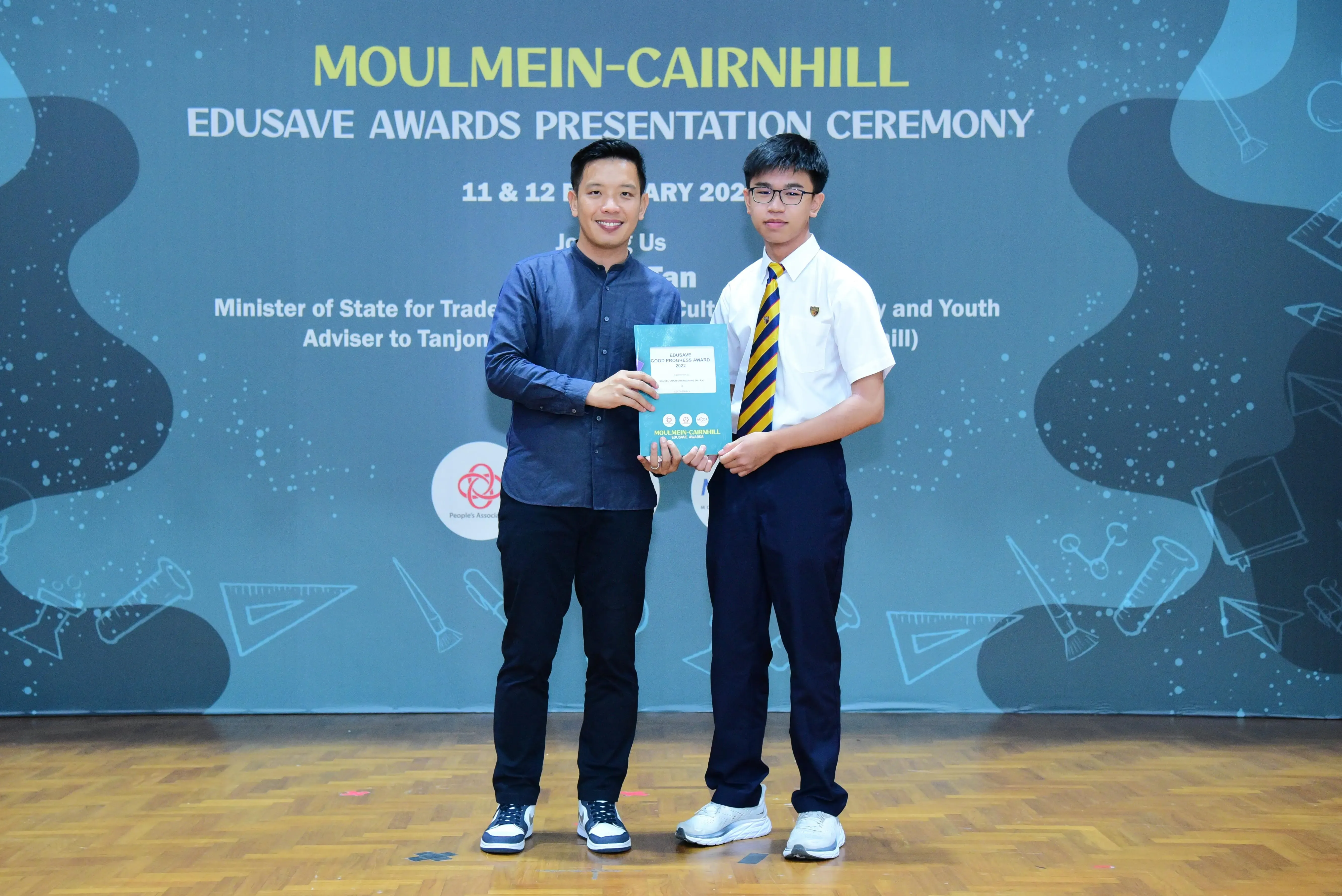 Two men holding a certificate at the Moulmein-Cairnhill Edusave Awards Presentation Ceremony backdrop.
