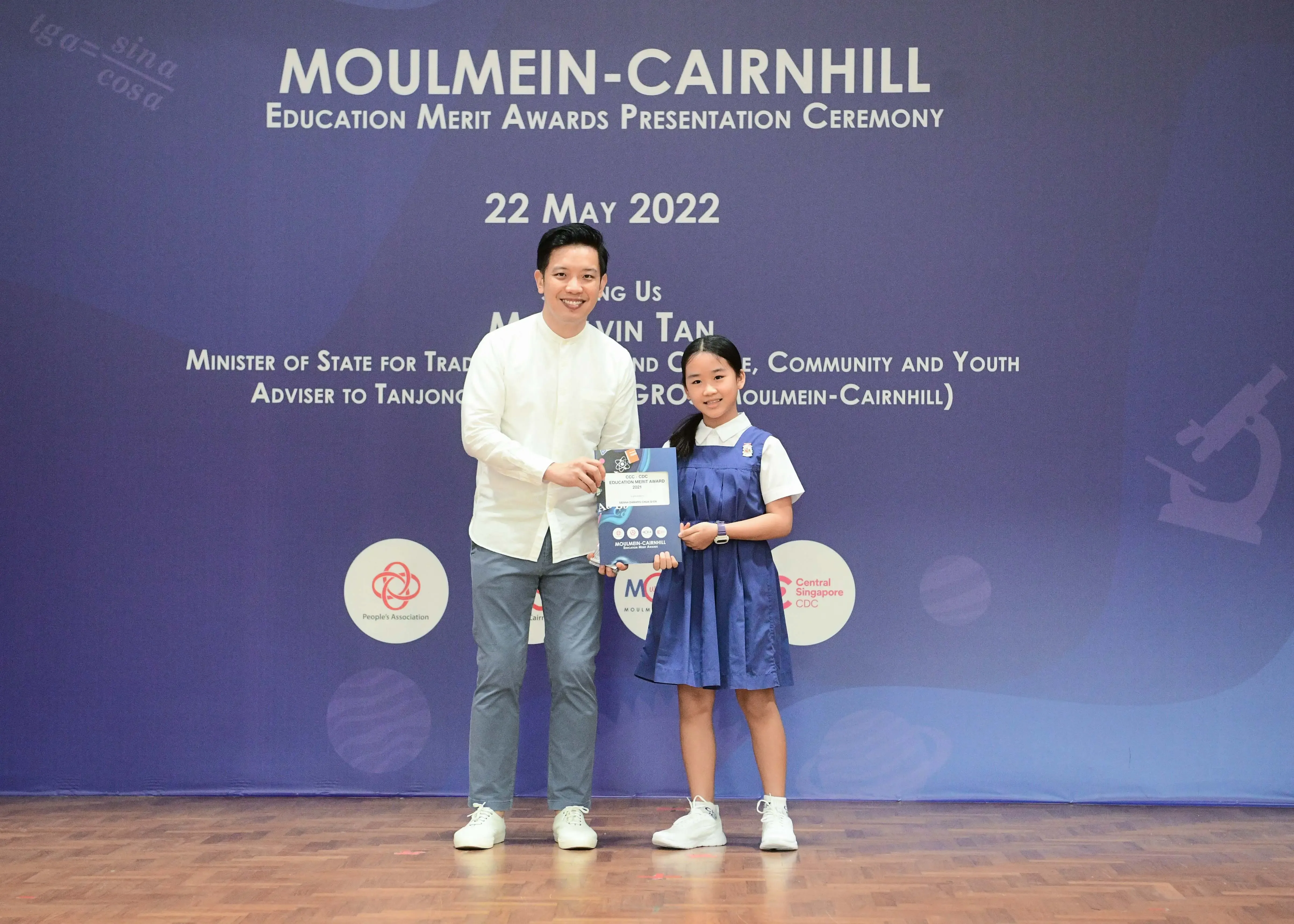 A man and a young girl in school uniform hold an award at the Moulmein-Cairnhill Education Merit Awards Ceremony, May 2022.