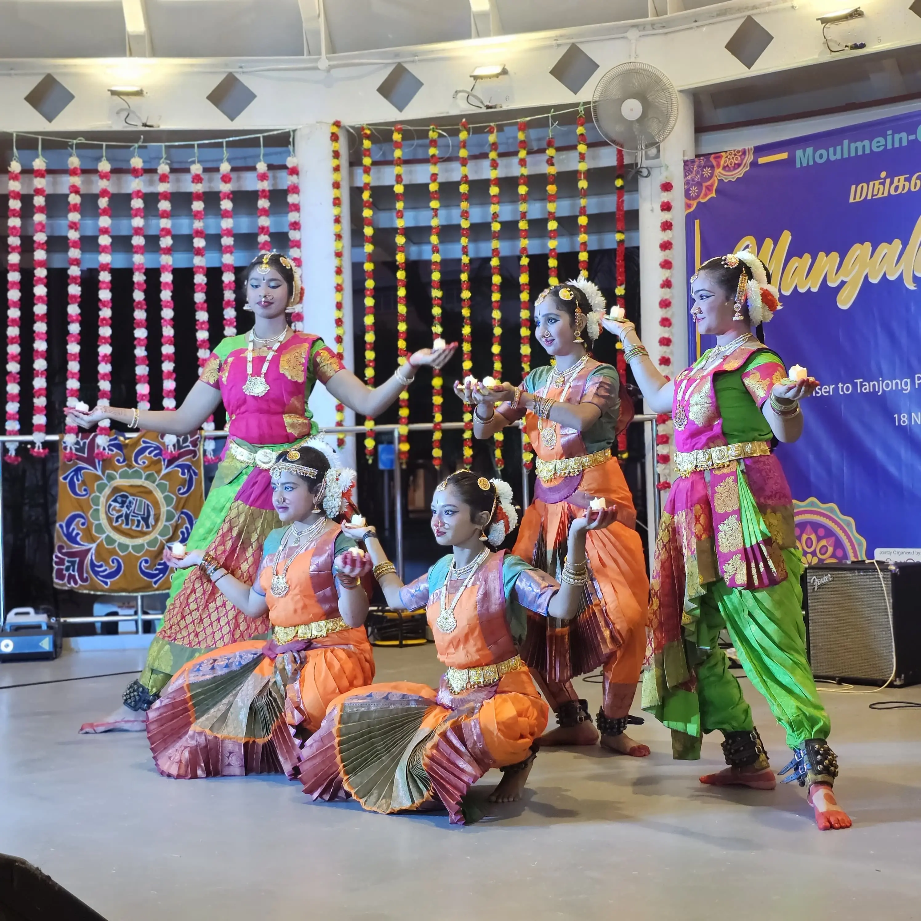 Five women in colorful traditional Indian attire perform a classical dance on stage, holding small oil lamps.