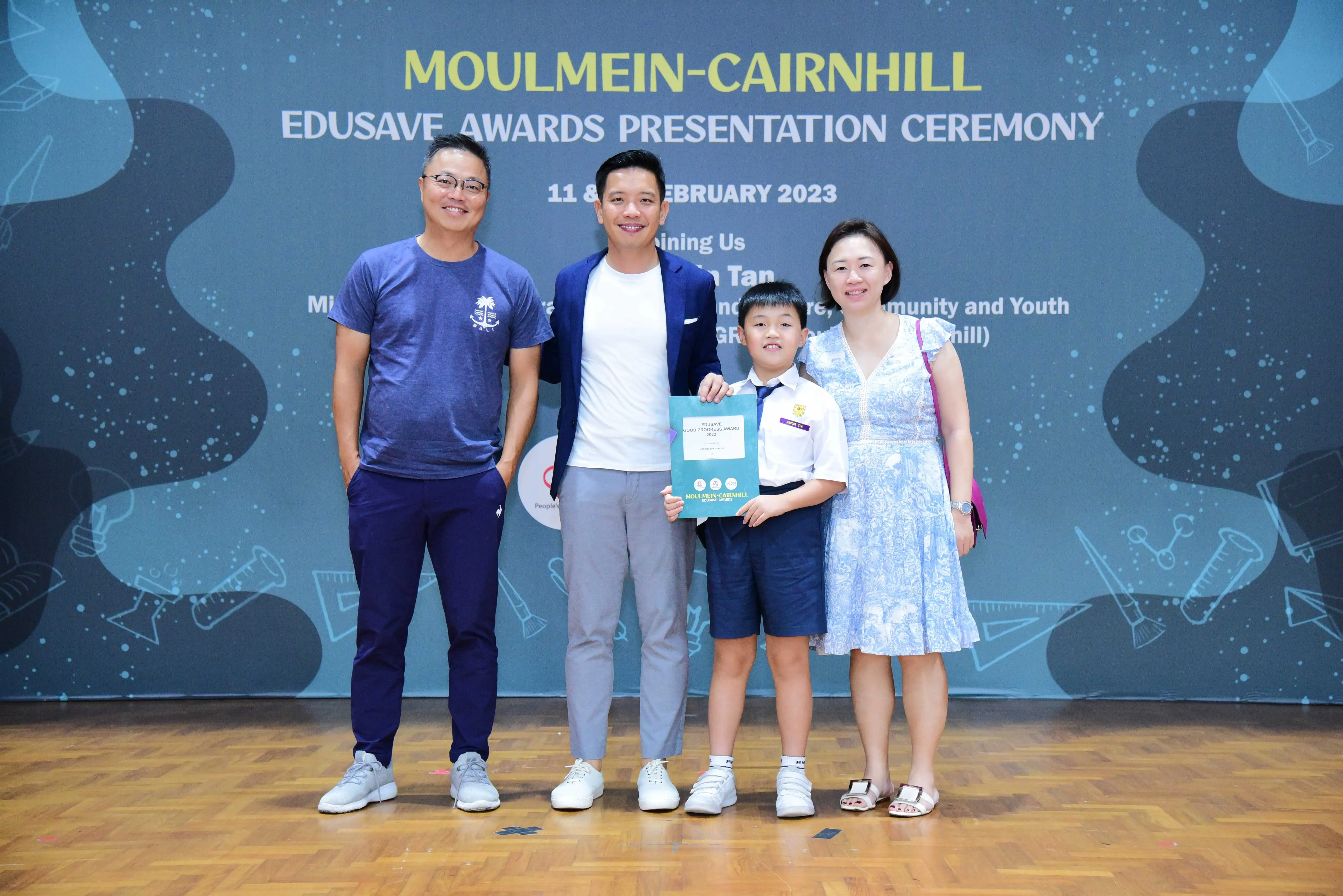 Four people pose at the Moulmein-Cairnhill Edusave Awards Presentation Ceremony, including a young boy holding a certificate.