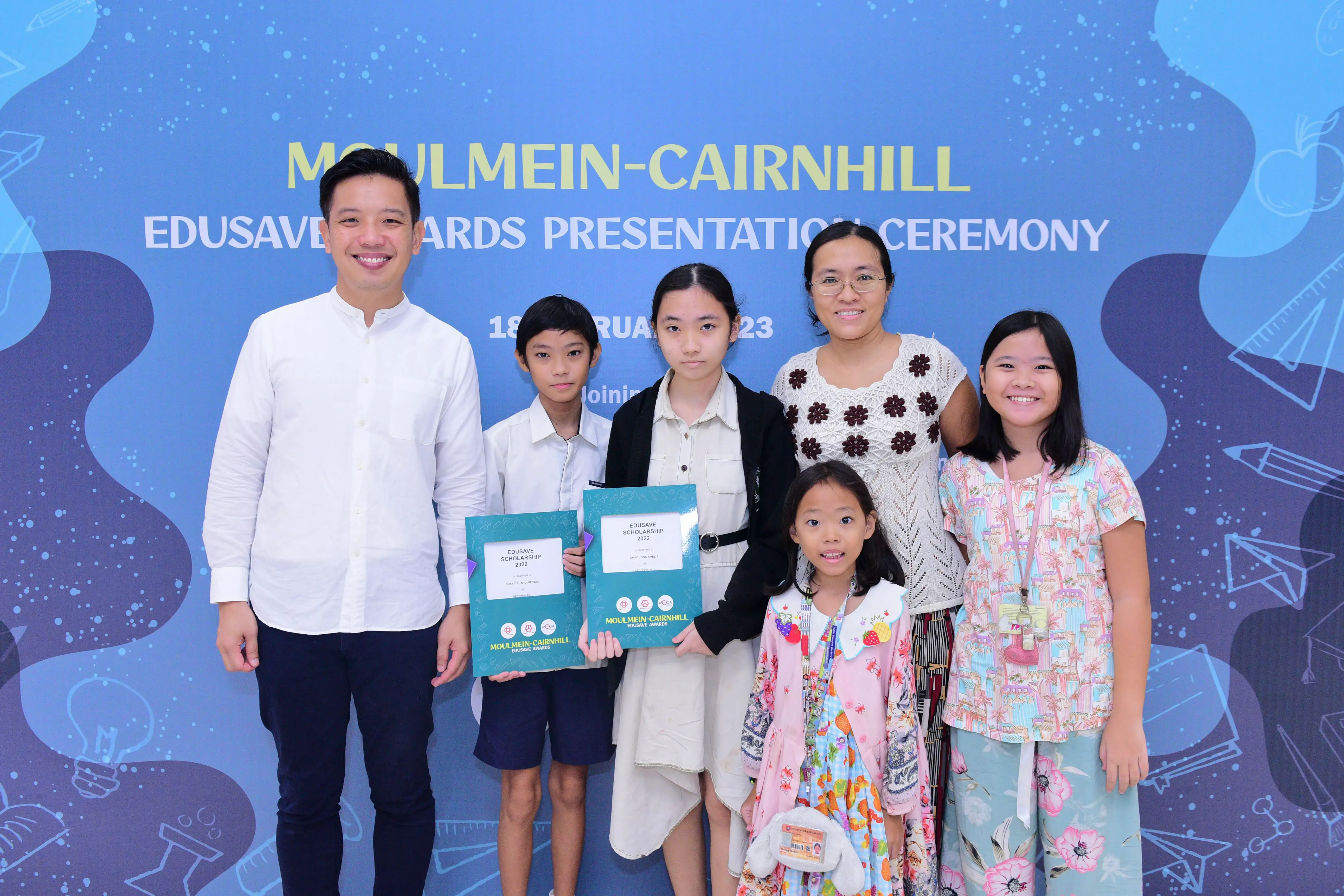 Six people pose in front of a Moulmein-Cairnhill Edusave Awards Presentation Ceremony banner, two holding teal award folders.
