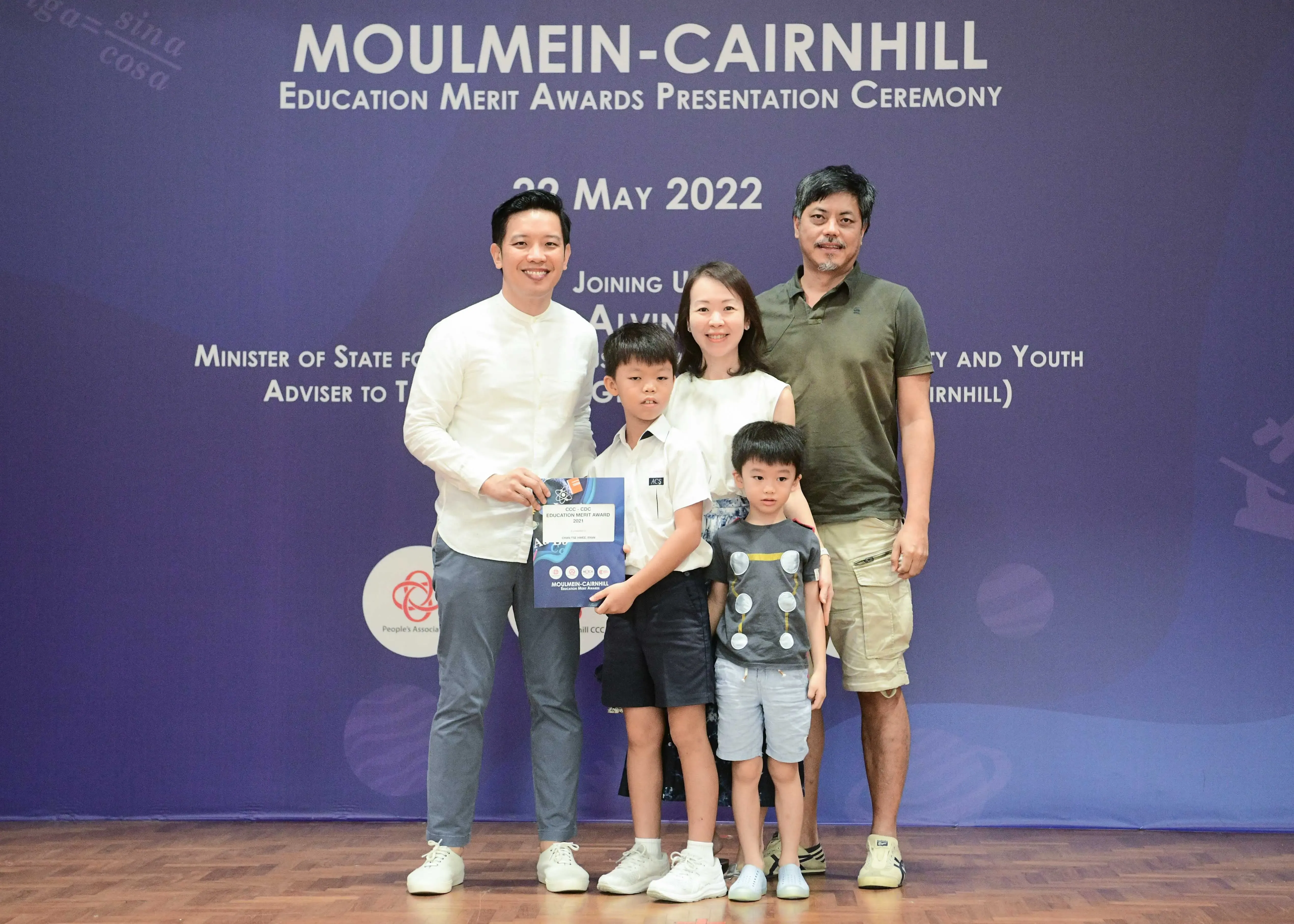 Five people pose before a Moulmein-Cairnhill Education Merit Awards banner dated 29 May 2022, with a boy holding a certificate.