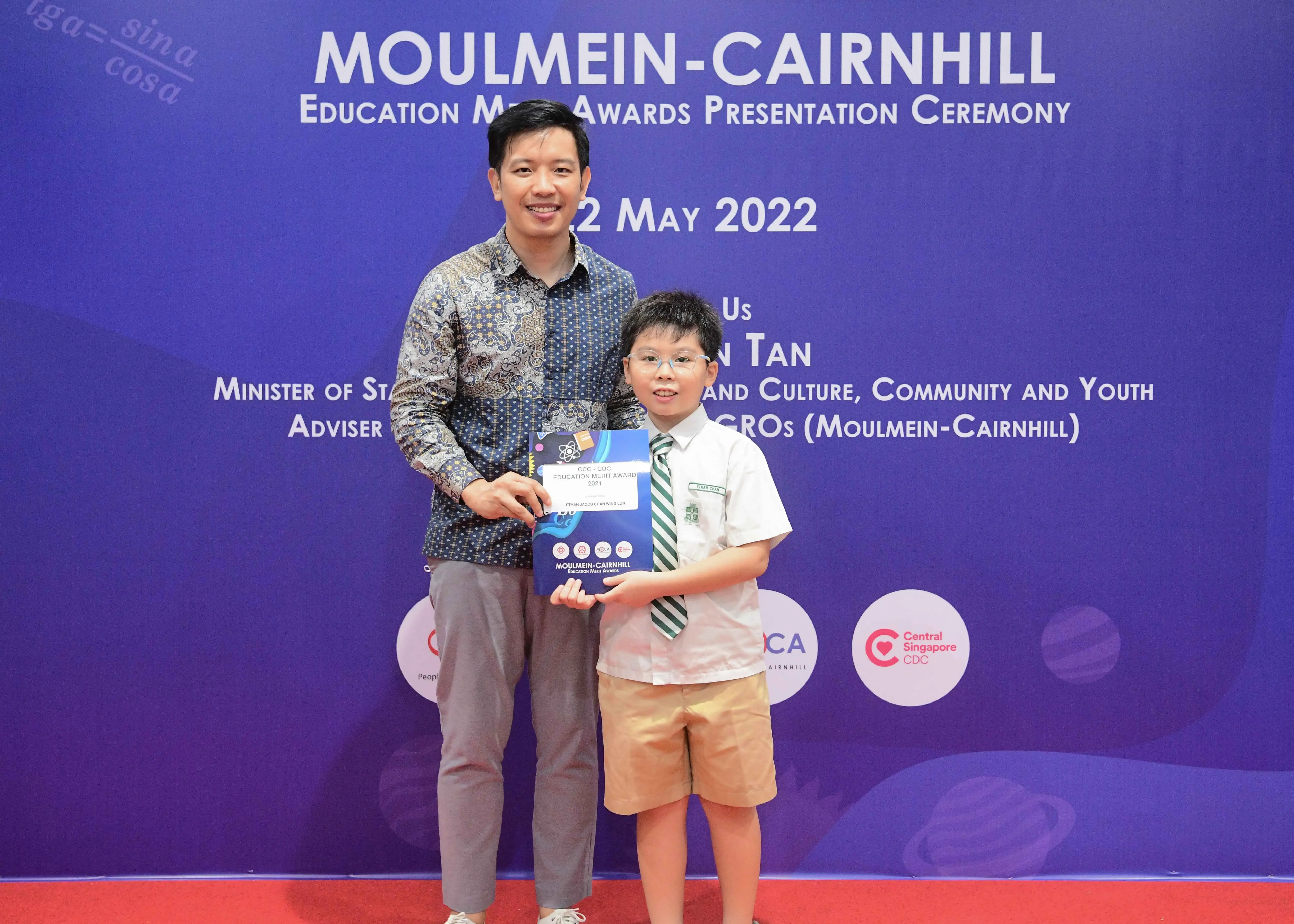 Adult man and young boy holding an award certificate at the Moulmein-Cairnhill Education Merit Awards Ceremony, May 2022.