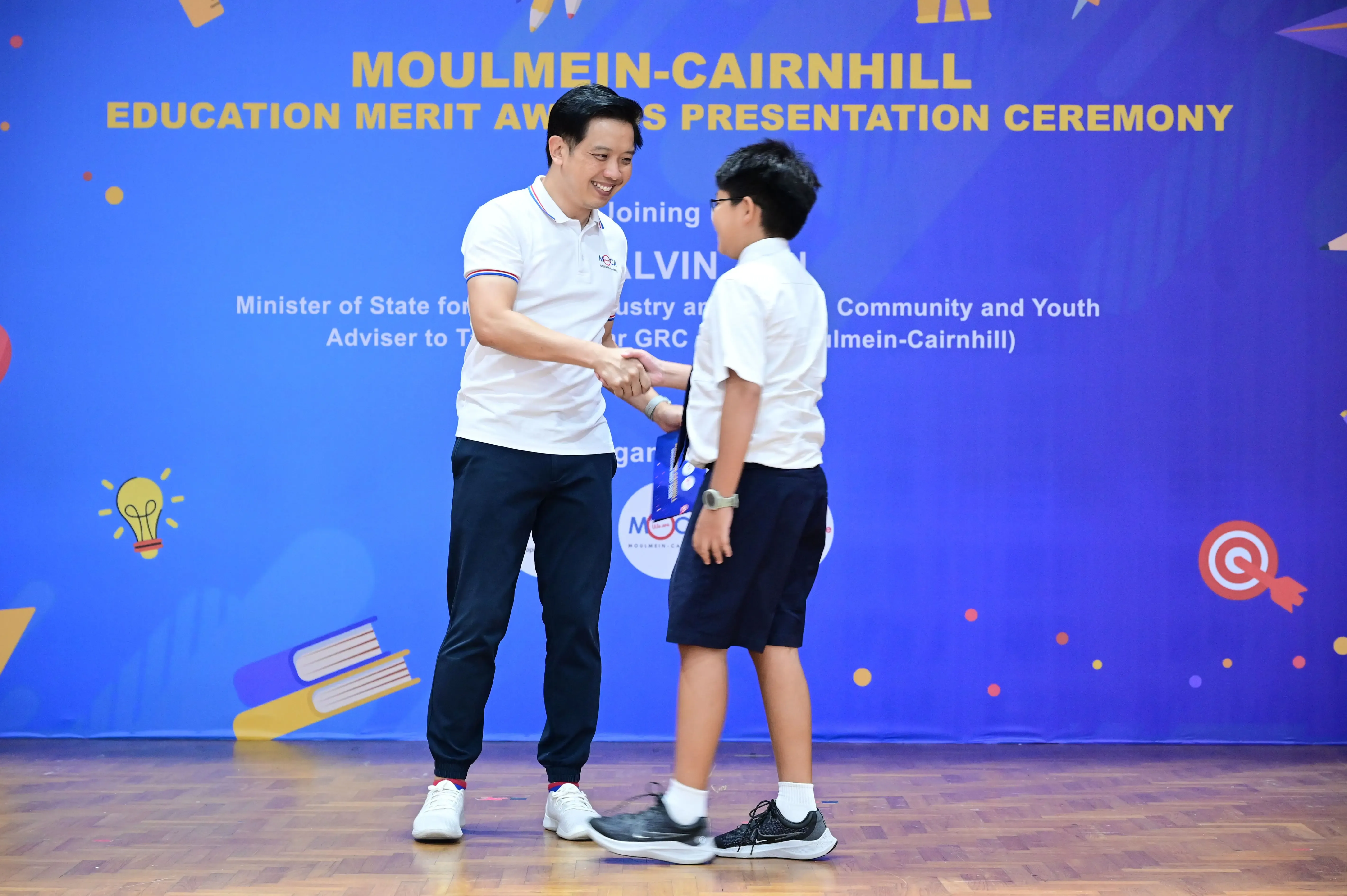 A man in a white polo shakes hands with a young boy at the Moulmein-Cairnhill Education Merit Awards ceremony.