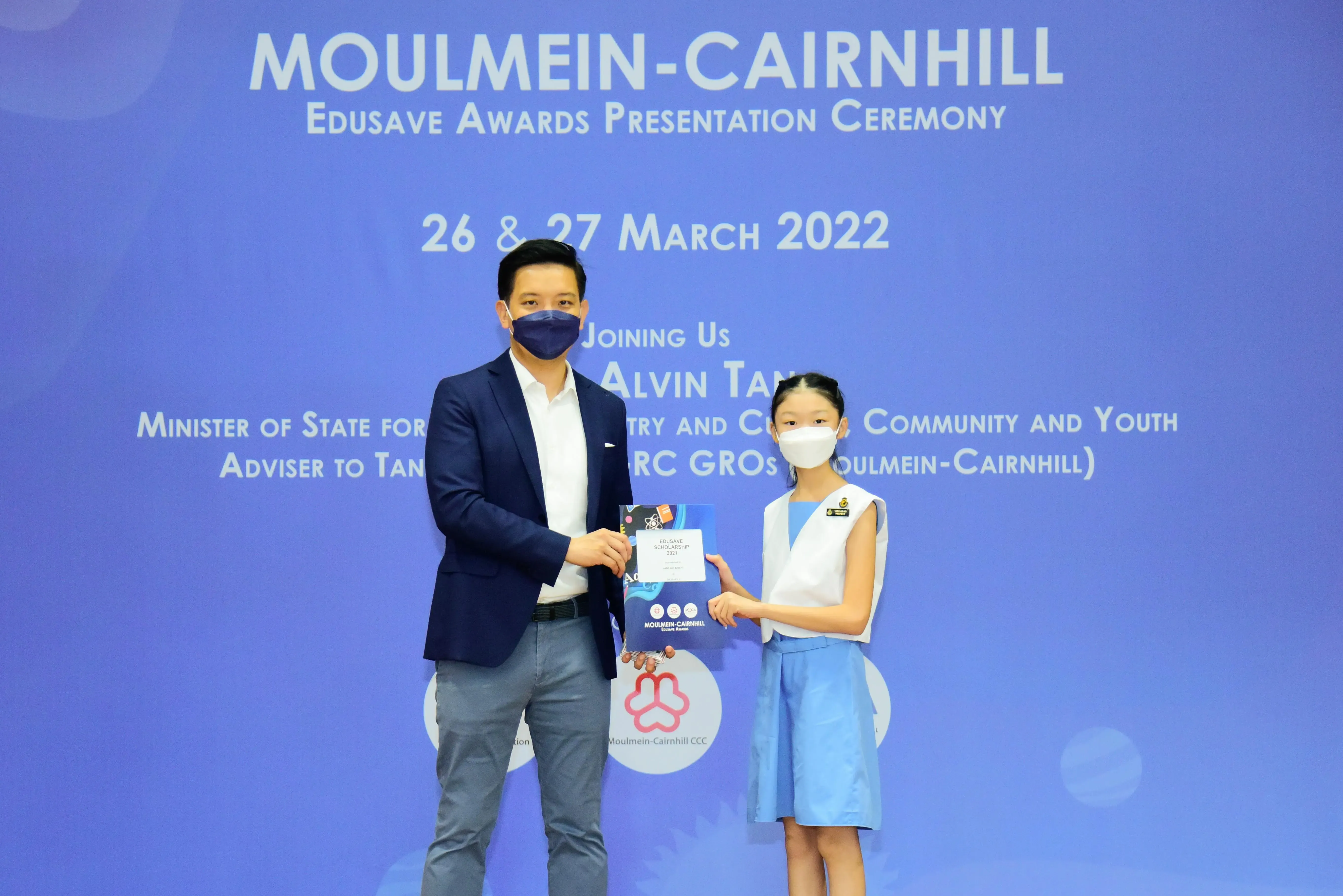 A masked man in a suit presents an Edusave award to a young girl at the Moulmein-Cairnhill ceremony on March 2022.