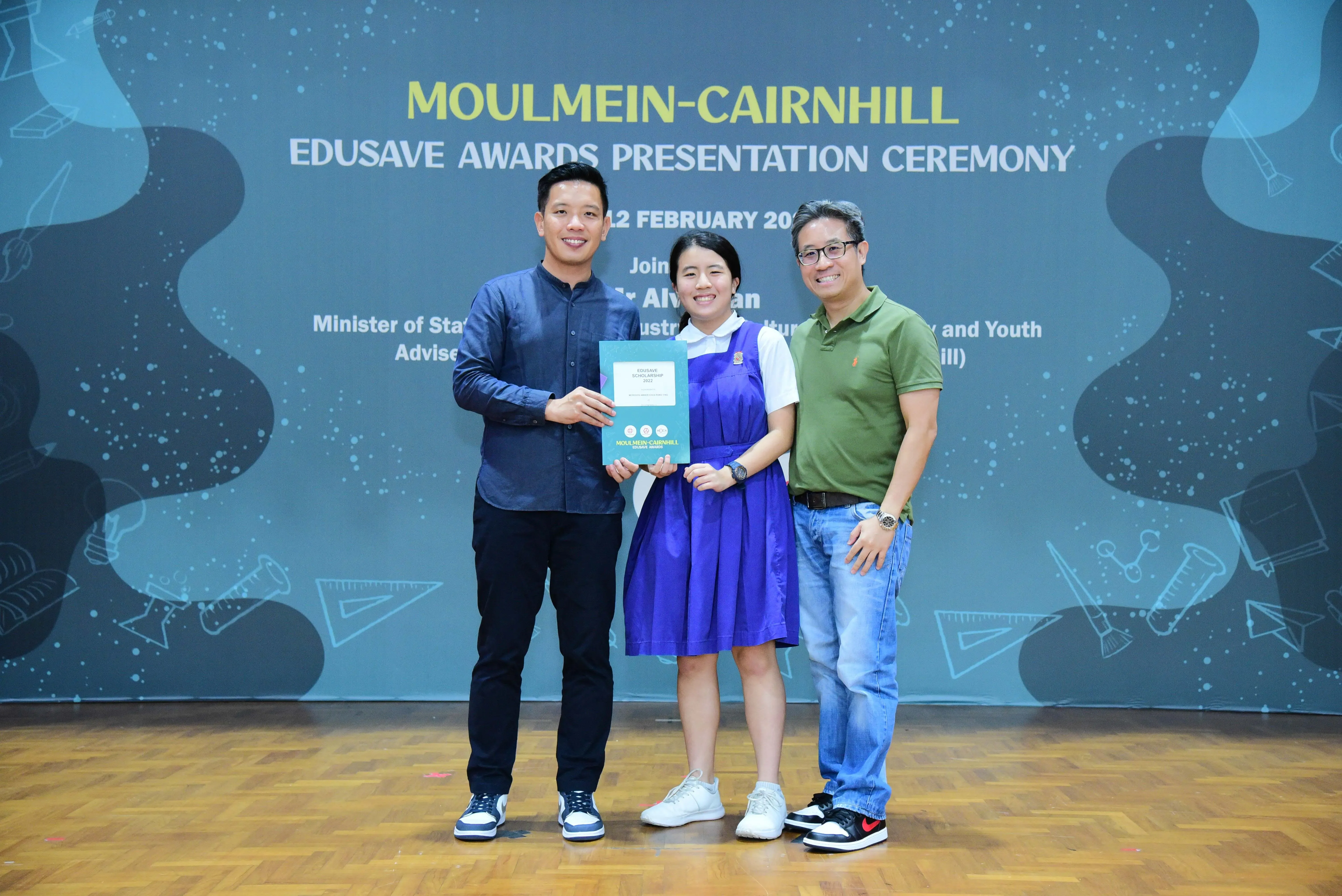 Three people pose with a certificate at the Moulmein-Cairnhill Edusave Awards Presentation Ceremony backdrop.