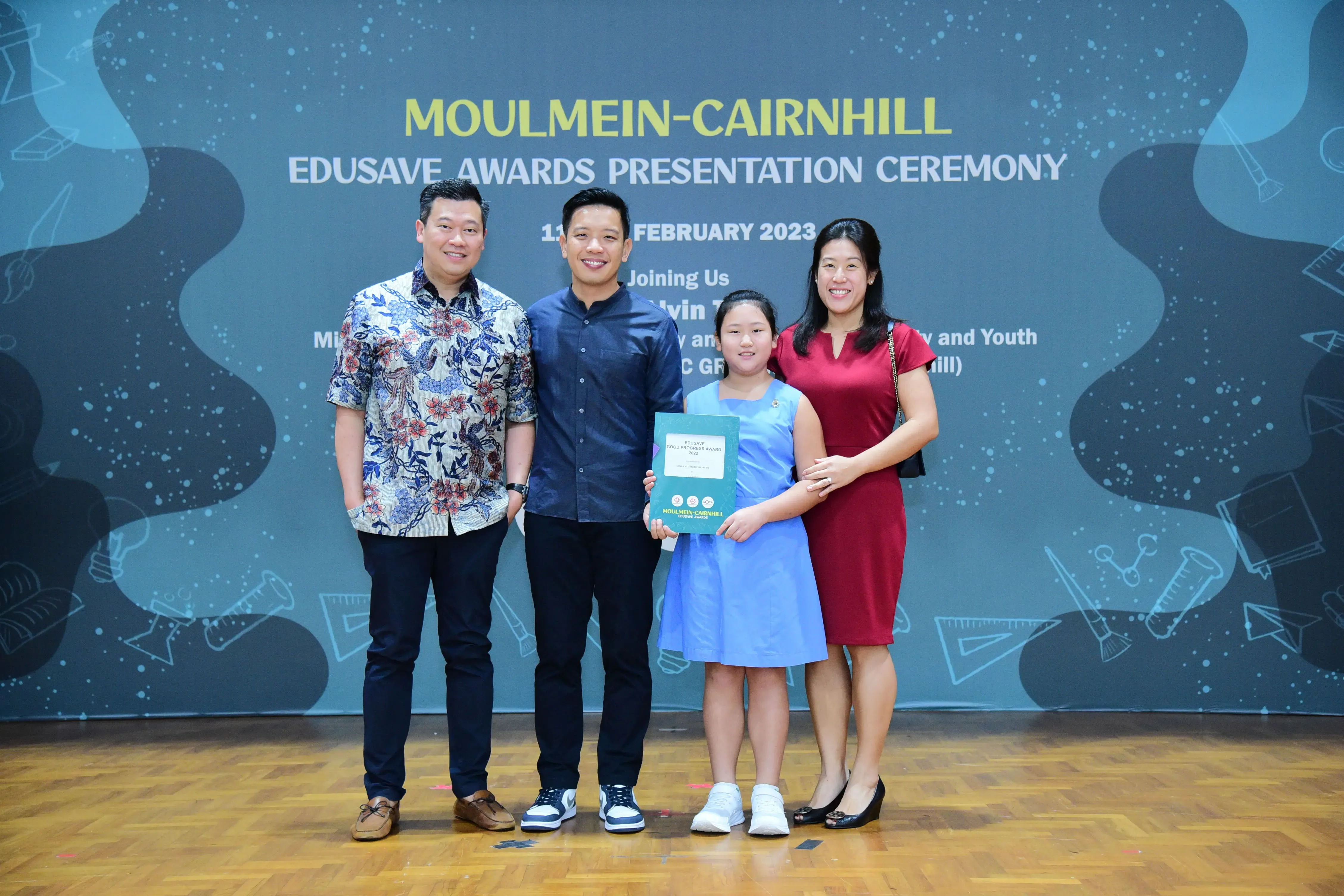 Four people pose before a Moulmein-Cairnhill Edusave Awards Presentation Ceremony banner; a girl holds a certificate.