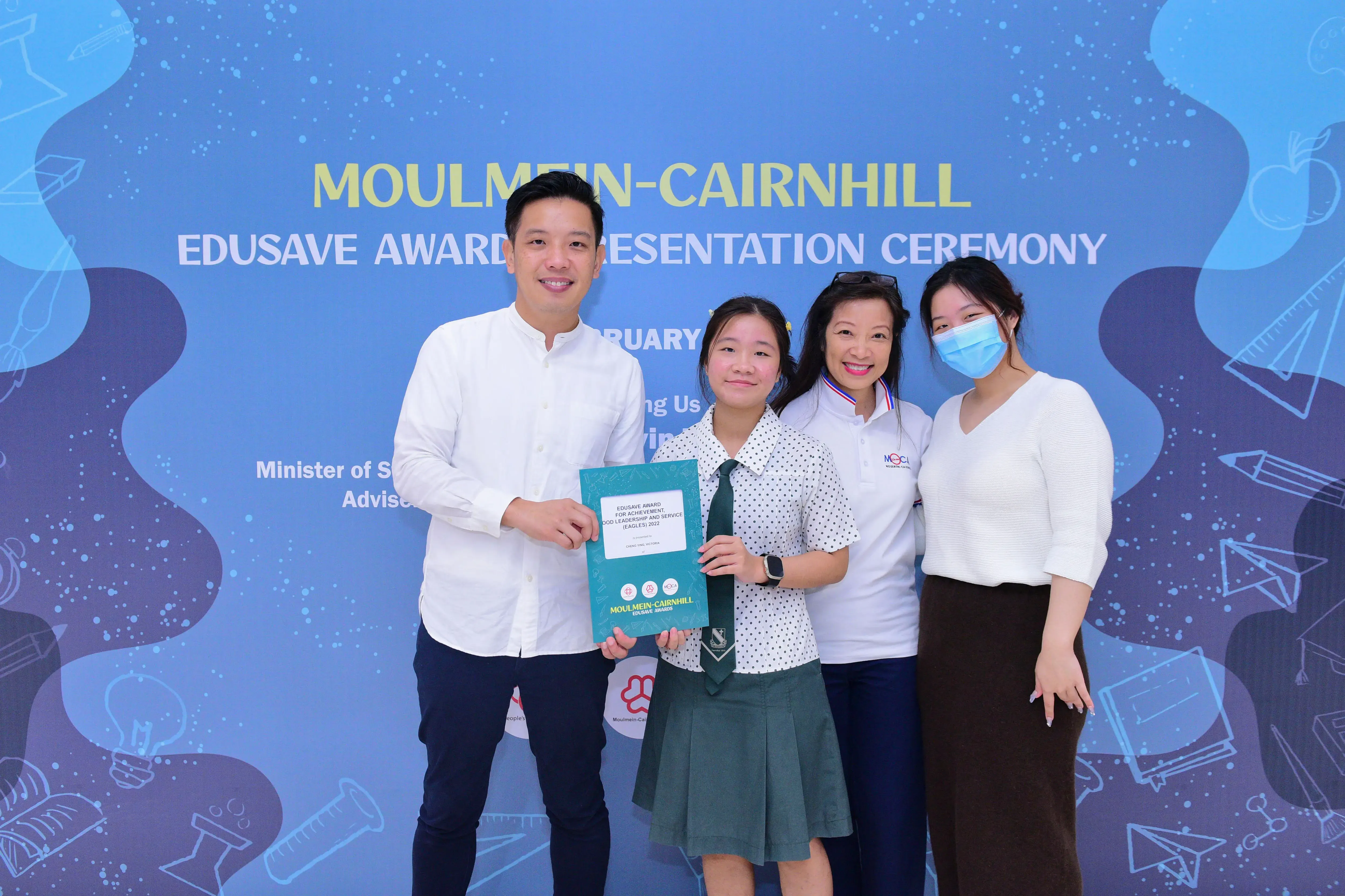 Four people pose at the Moulmein-Cairnhill Edusave Award Presentation Ceremony, with a student holding a teal award folder.