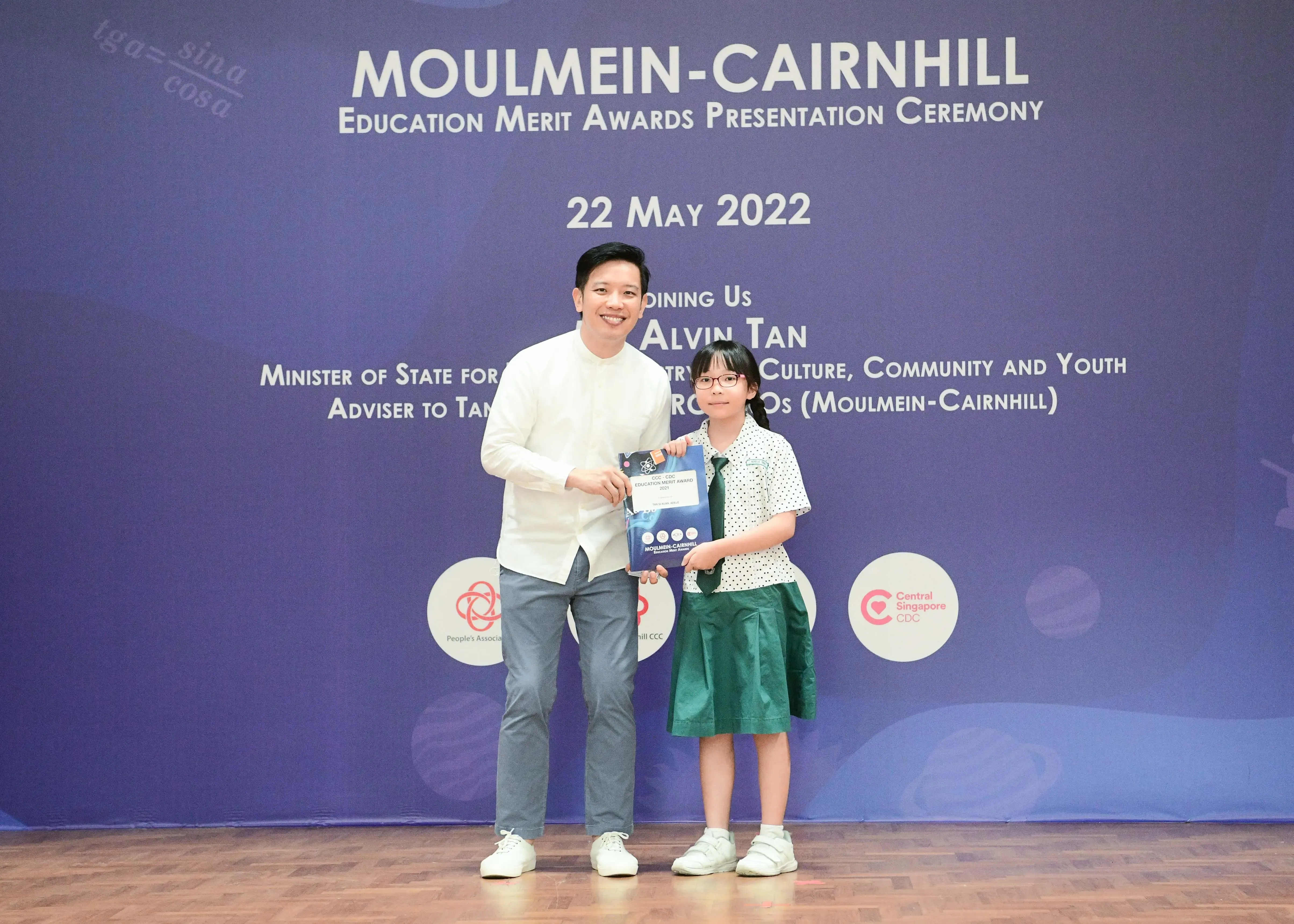 A man and a young girl in school uniform hold an award together at the Moulmein-Cairnhill Education Merit Awards ceremony.