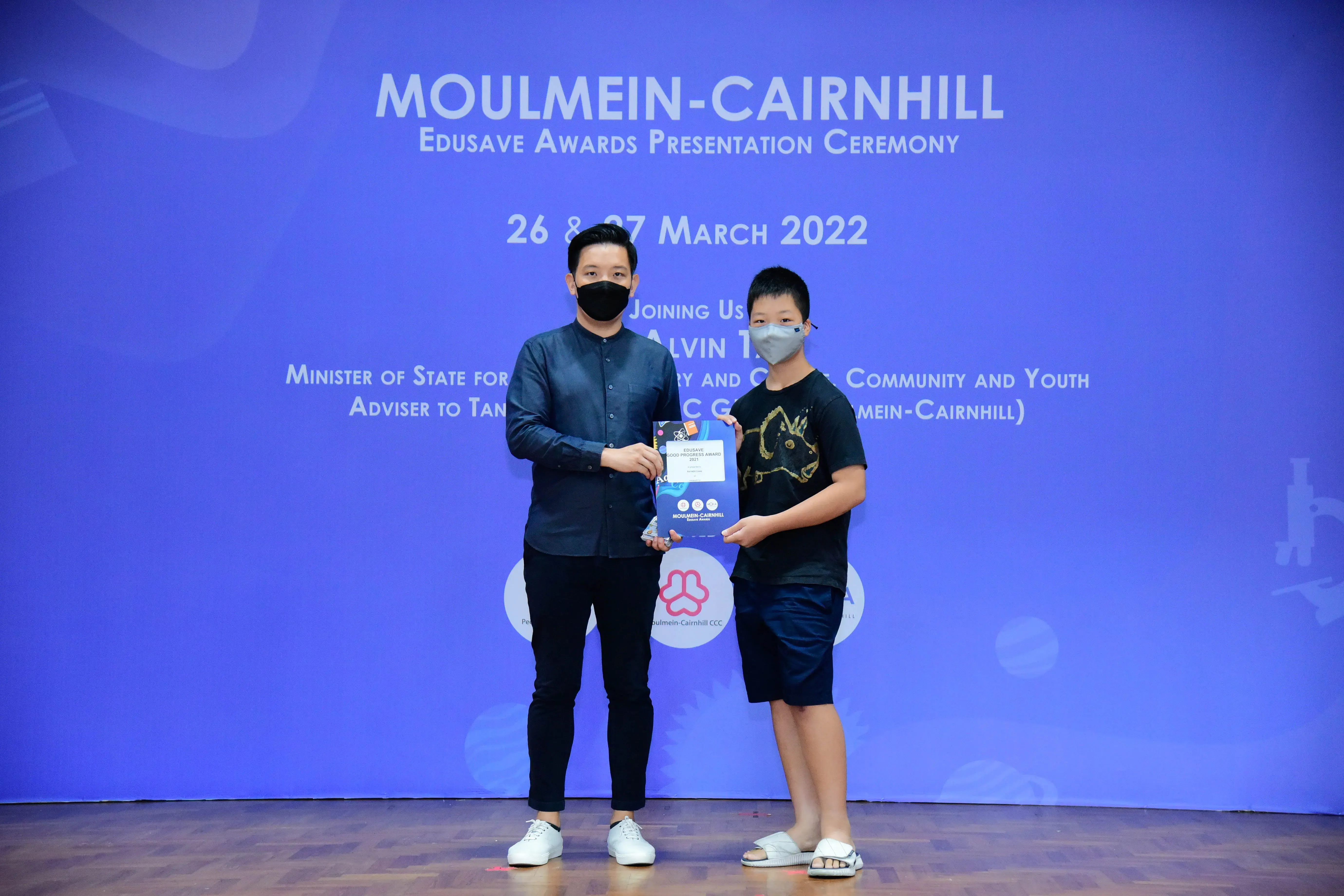 An adult and a young boy wearing masks hold a certificate at the Moulmein-Cairnhill Edusave Awards 2022.