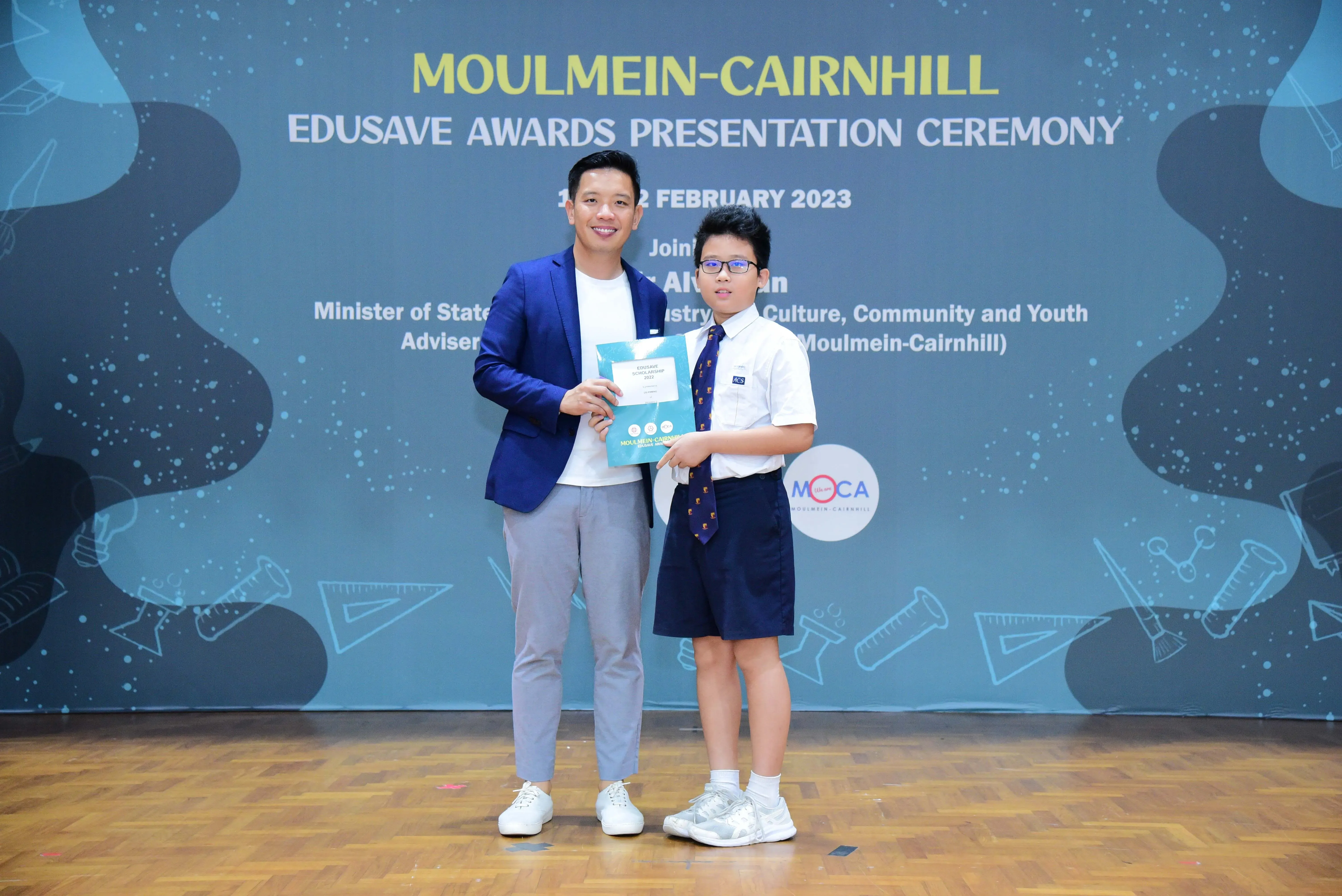 An adult in a blue blazer and a young student in uniform hold an award at the Moulmein-Cairnhill Edusave Awards Ceremony 2023.