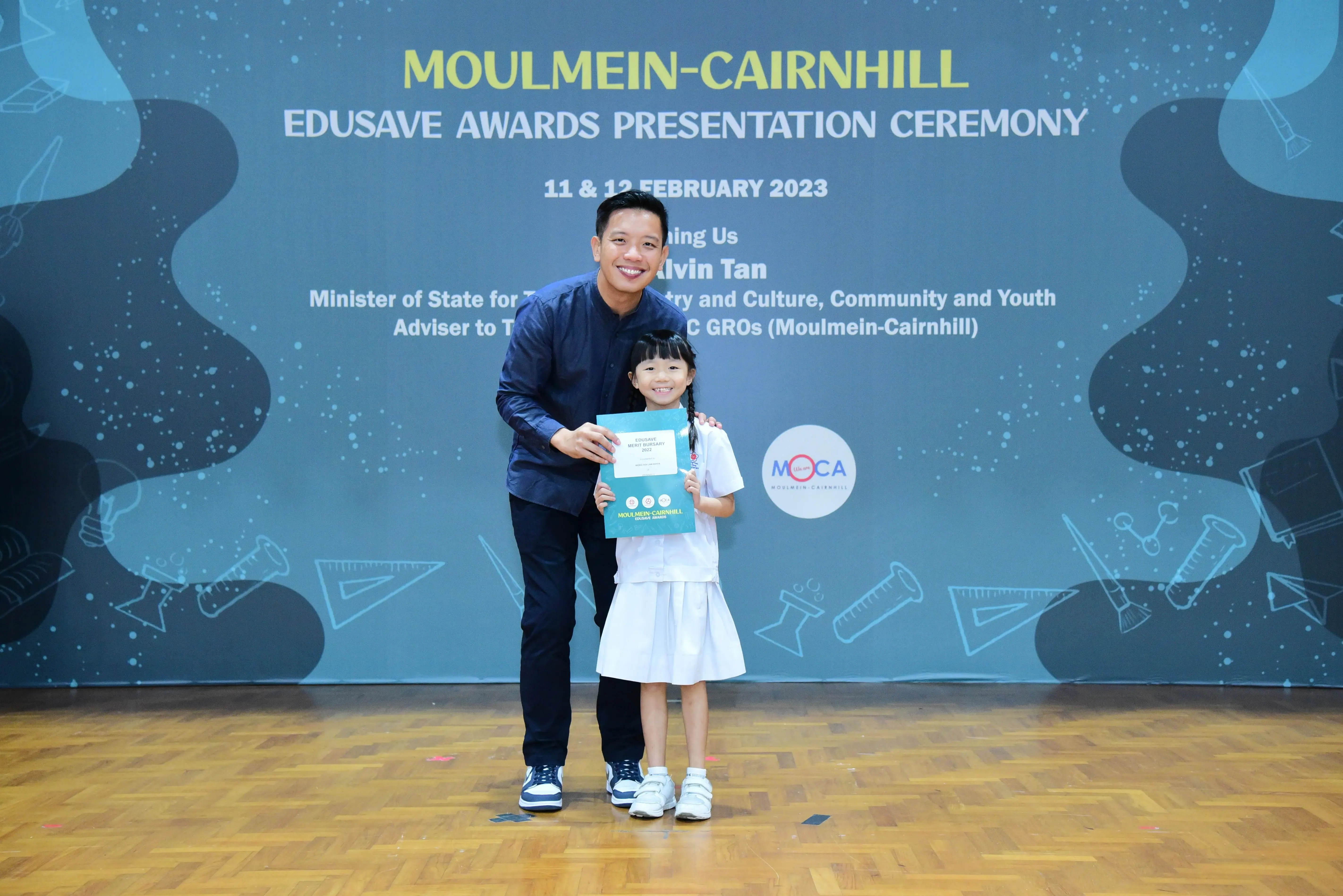 A man and a young girl in school uniform hold a certificate at the Moulmein-Cairnhill Edusave Awards Ceremony 2023.