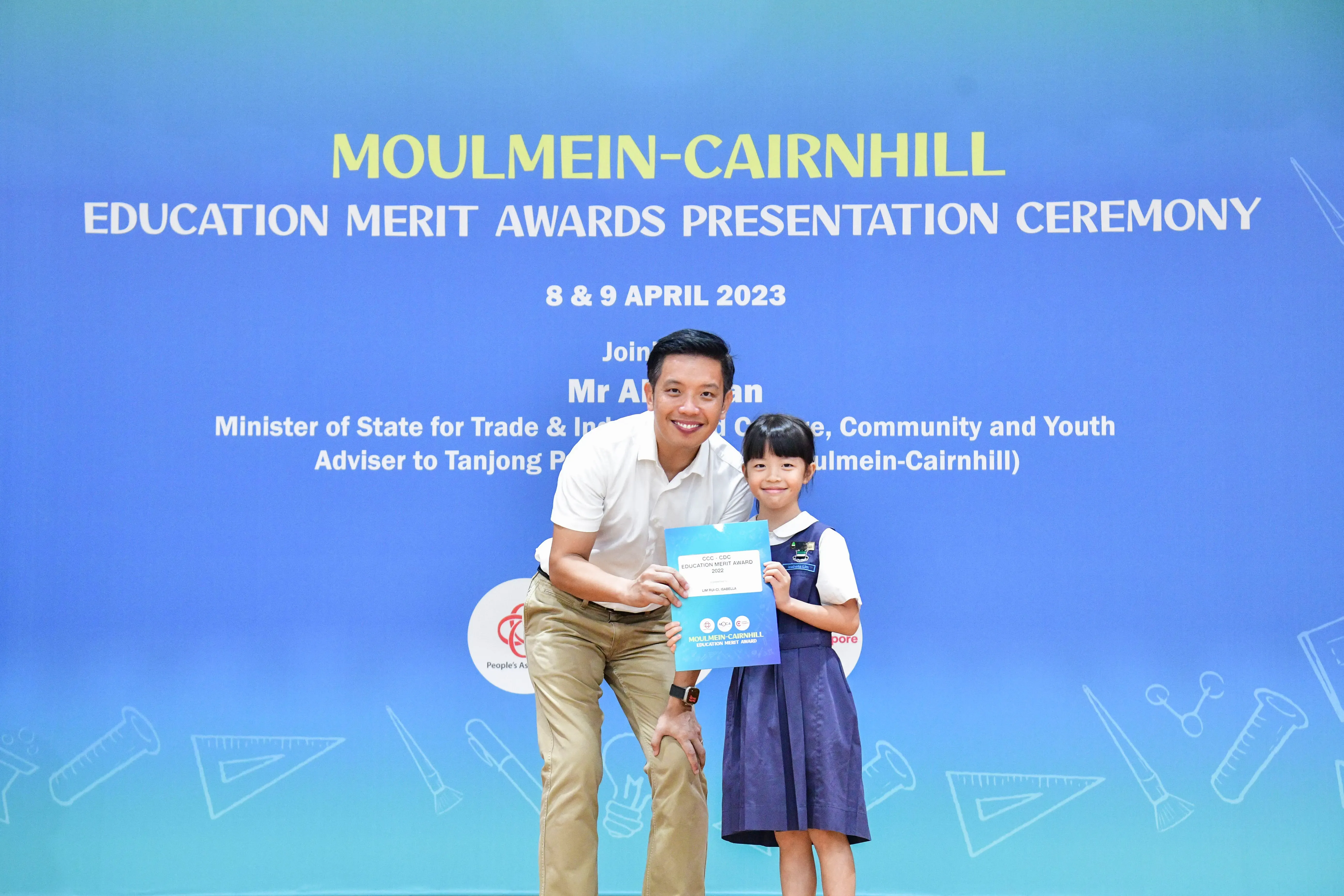 A man and a young girl hold an award certificate in front of a Moulmein-Cairnhill Education Merit Awards banner.