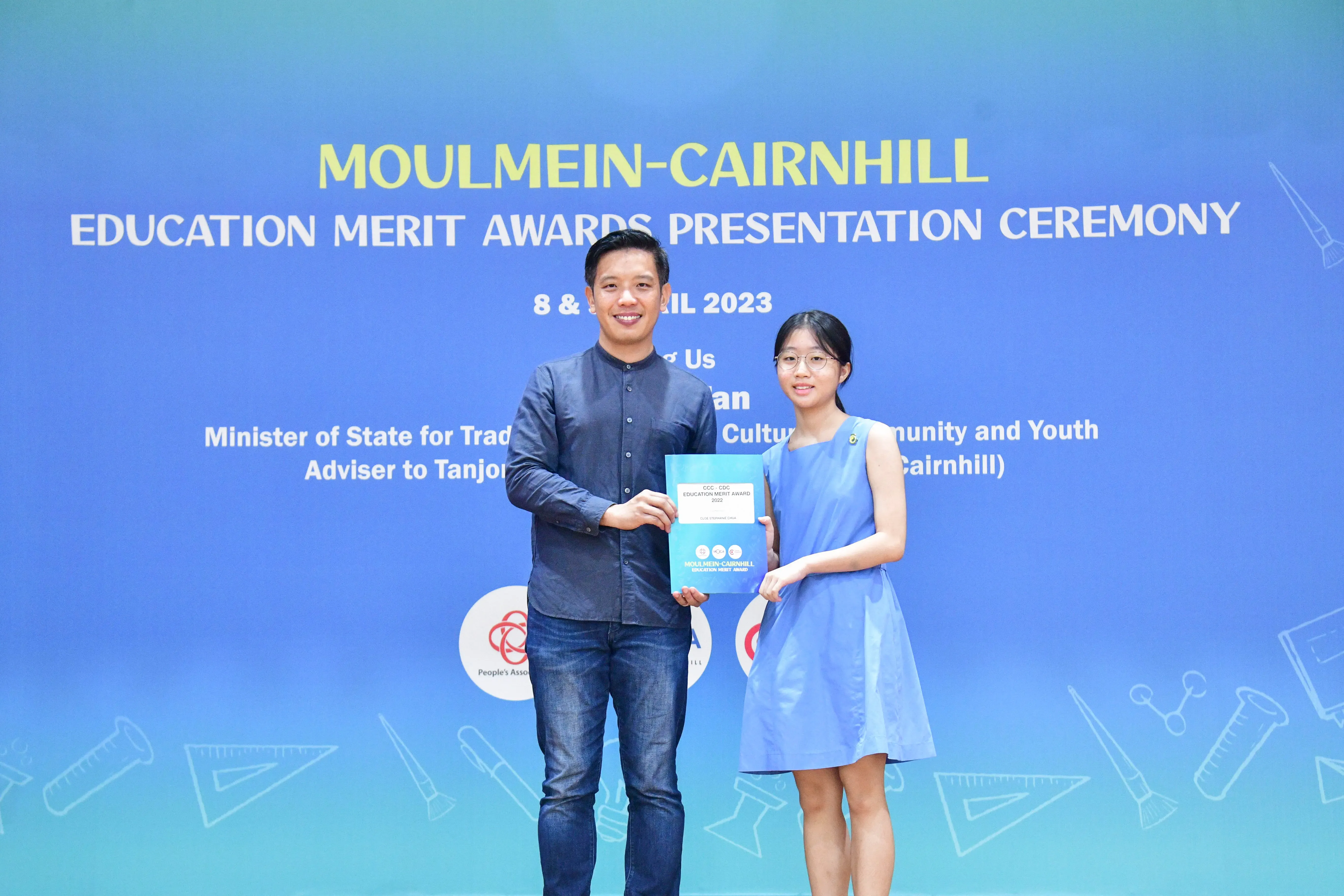 Two people hold a certificate together in front of a Moulmein-Cairnhill Education Merit Awards 2023 banner.