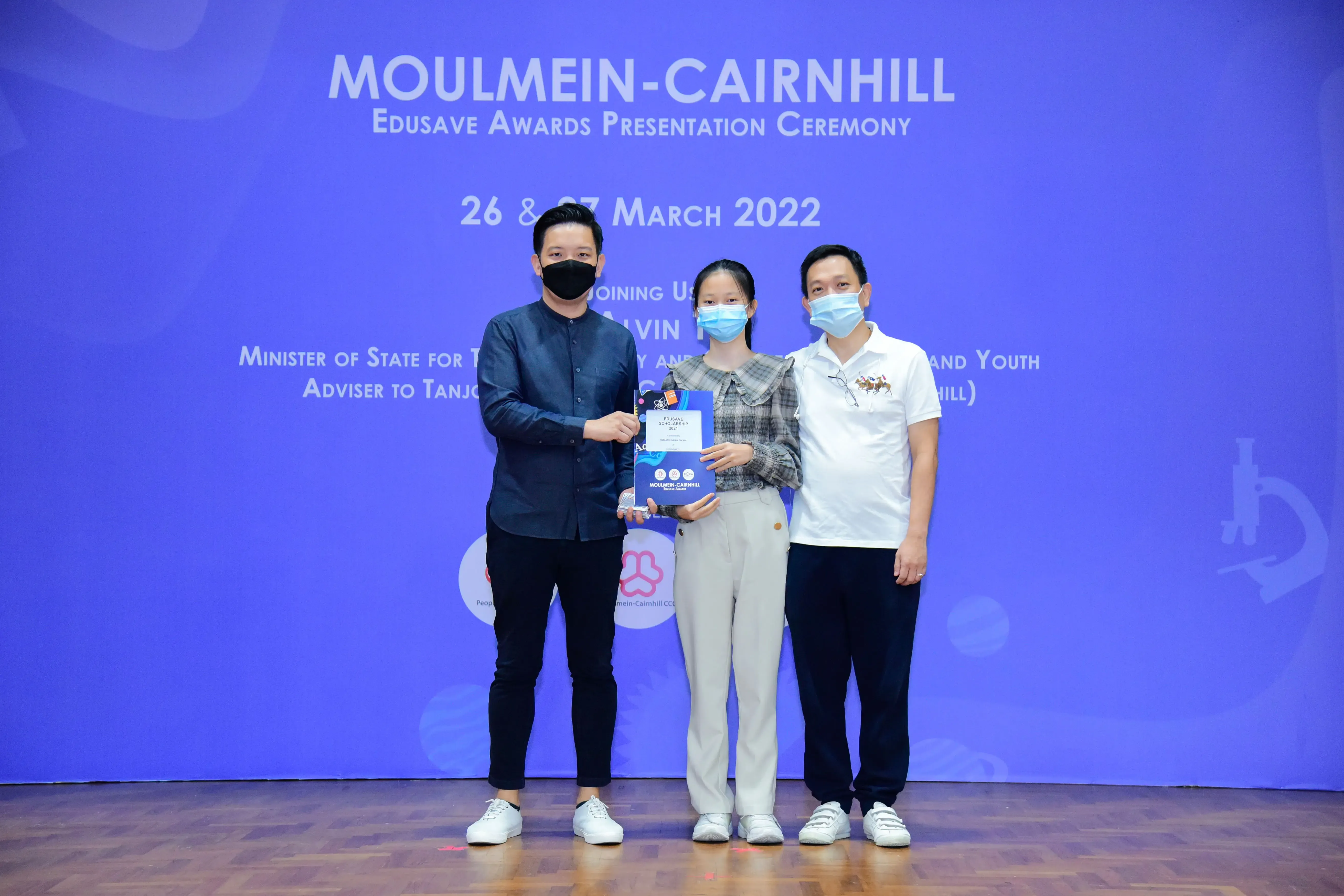 Three masked people pose on stage at the Moulmein-Cairnhill Edusave Awards Presentation Ceremony, March 2022.