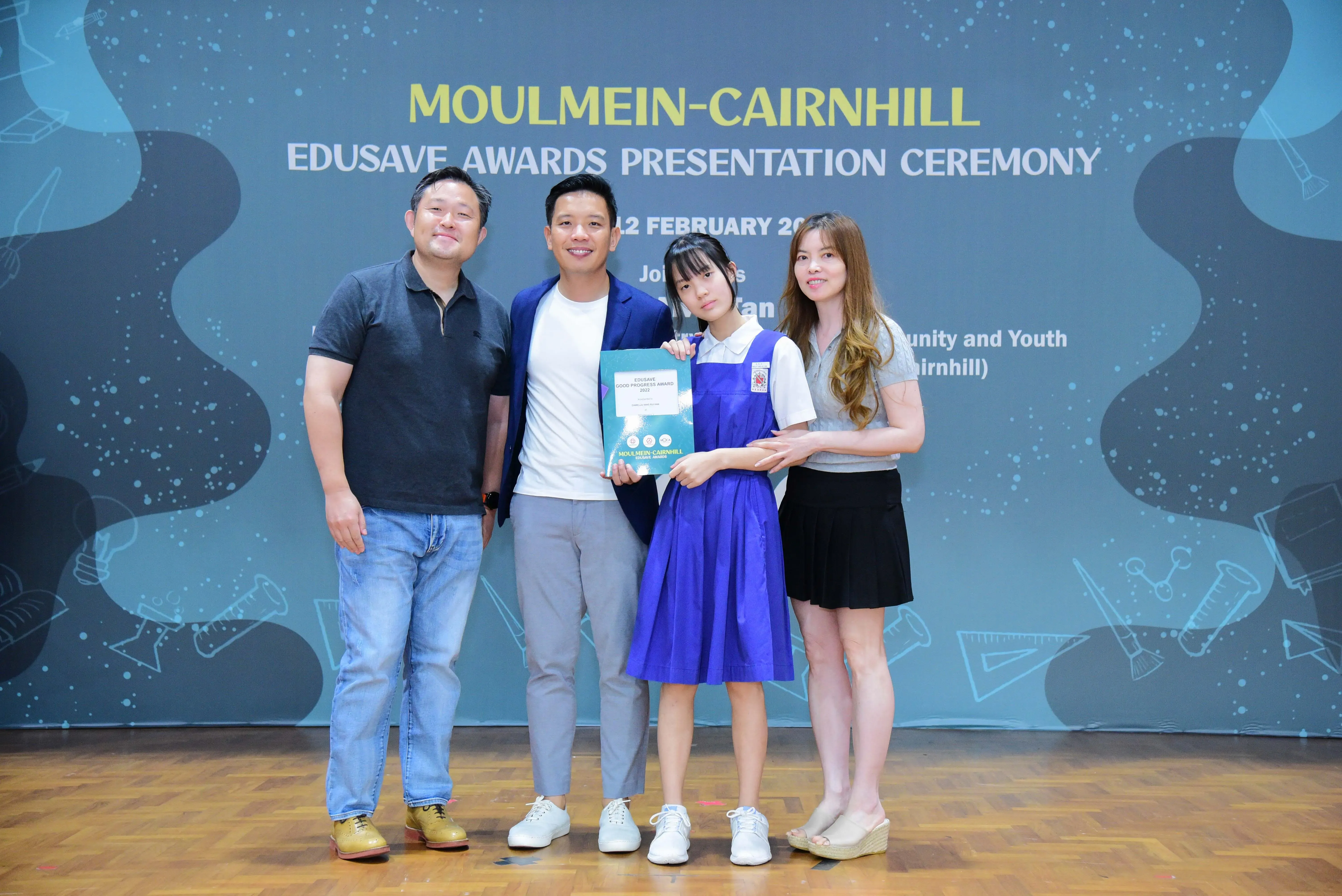 Four people pose at the Moulmein-Cairnhill Edusave Awards Presentation Ceremony, with a student holding an award certificate.