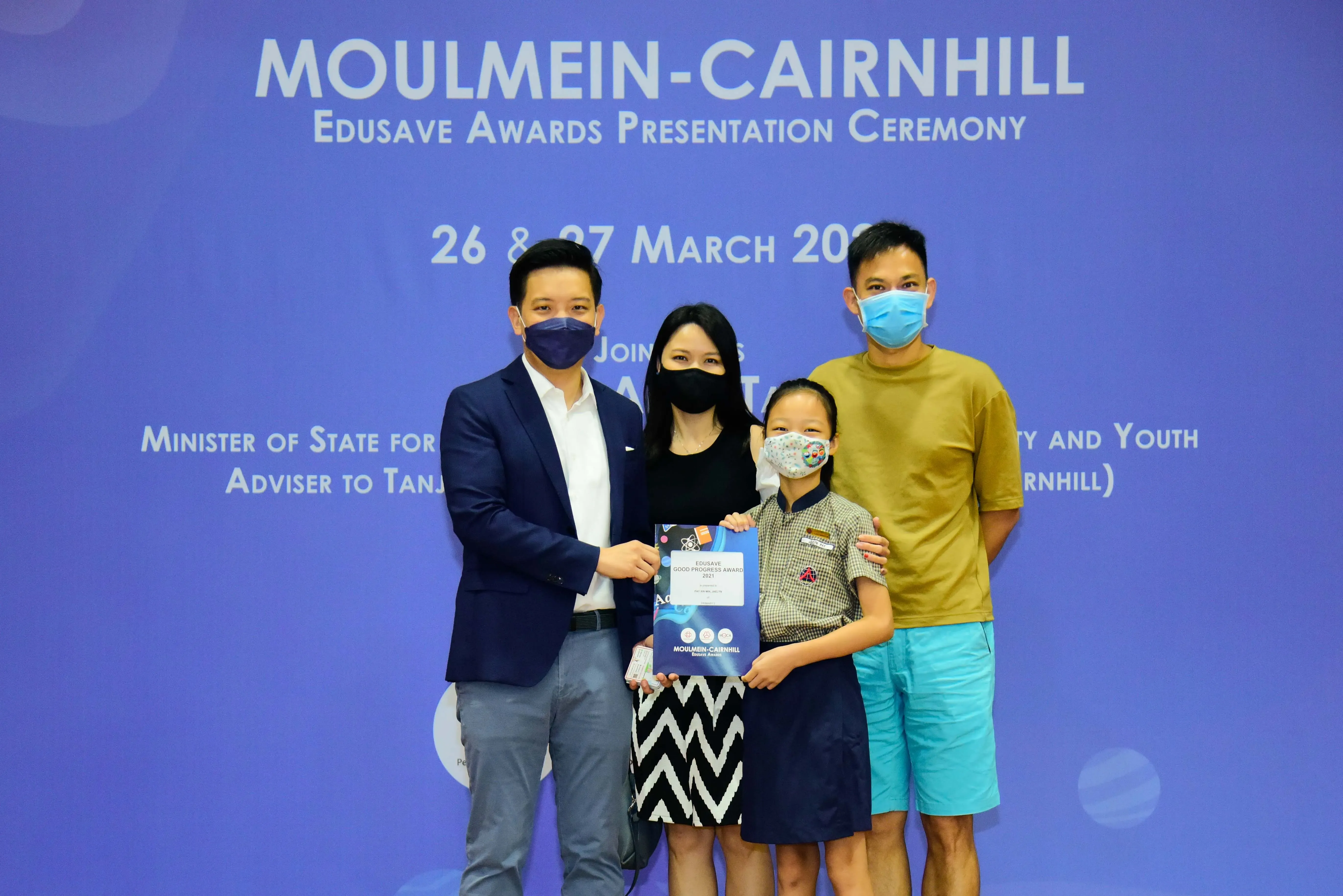 Four masked people pose at the Moulmein-Cairnhill Edusave Awards Presentation Ceremony, with a child holding an award.