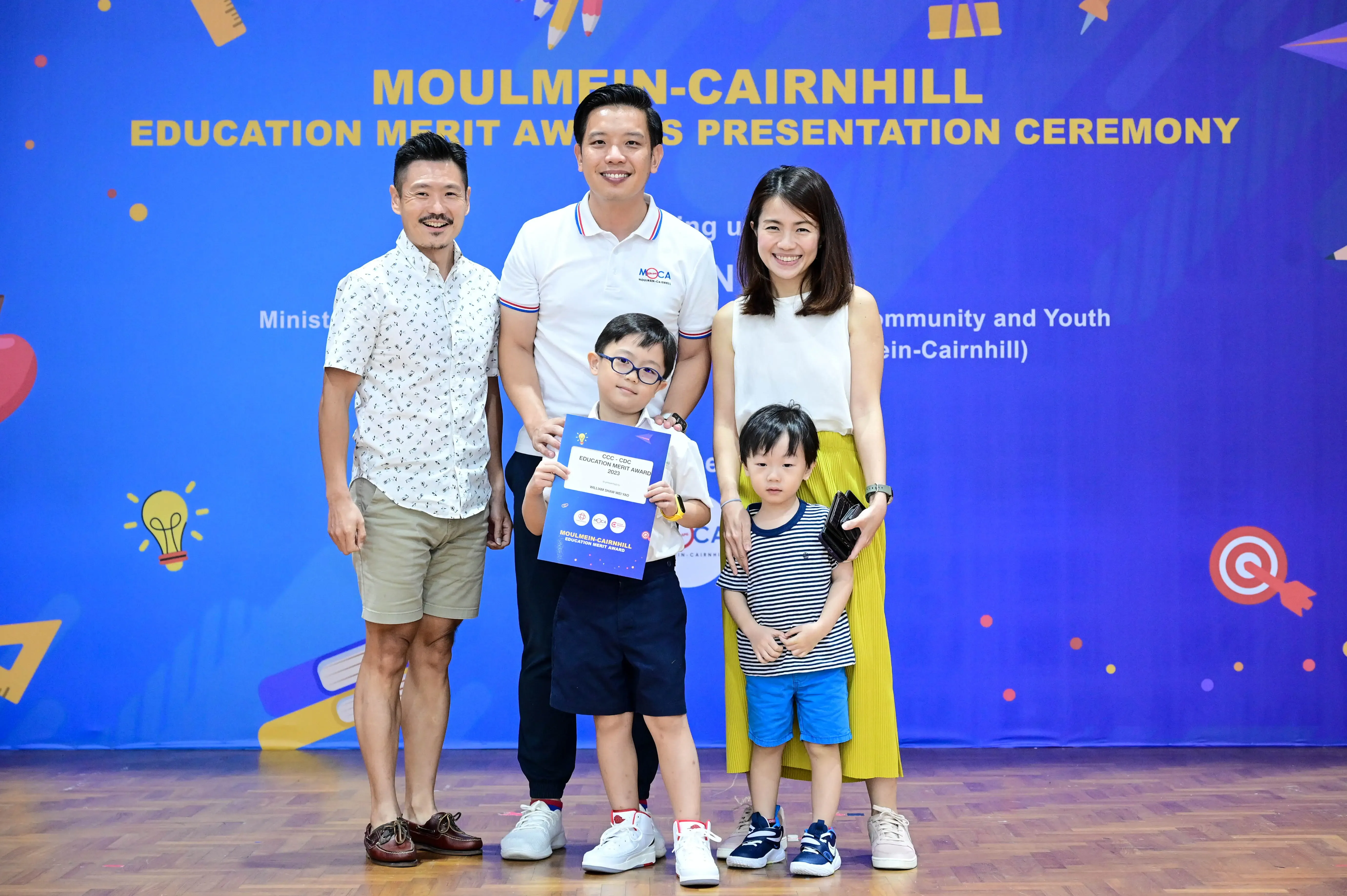 Three adults and two young boys pose at the Moulmein-Cairnhill Education Merit Awards Presentation Ceremony, one boy holding a certificate.