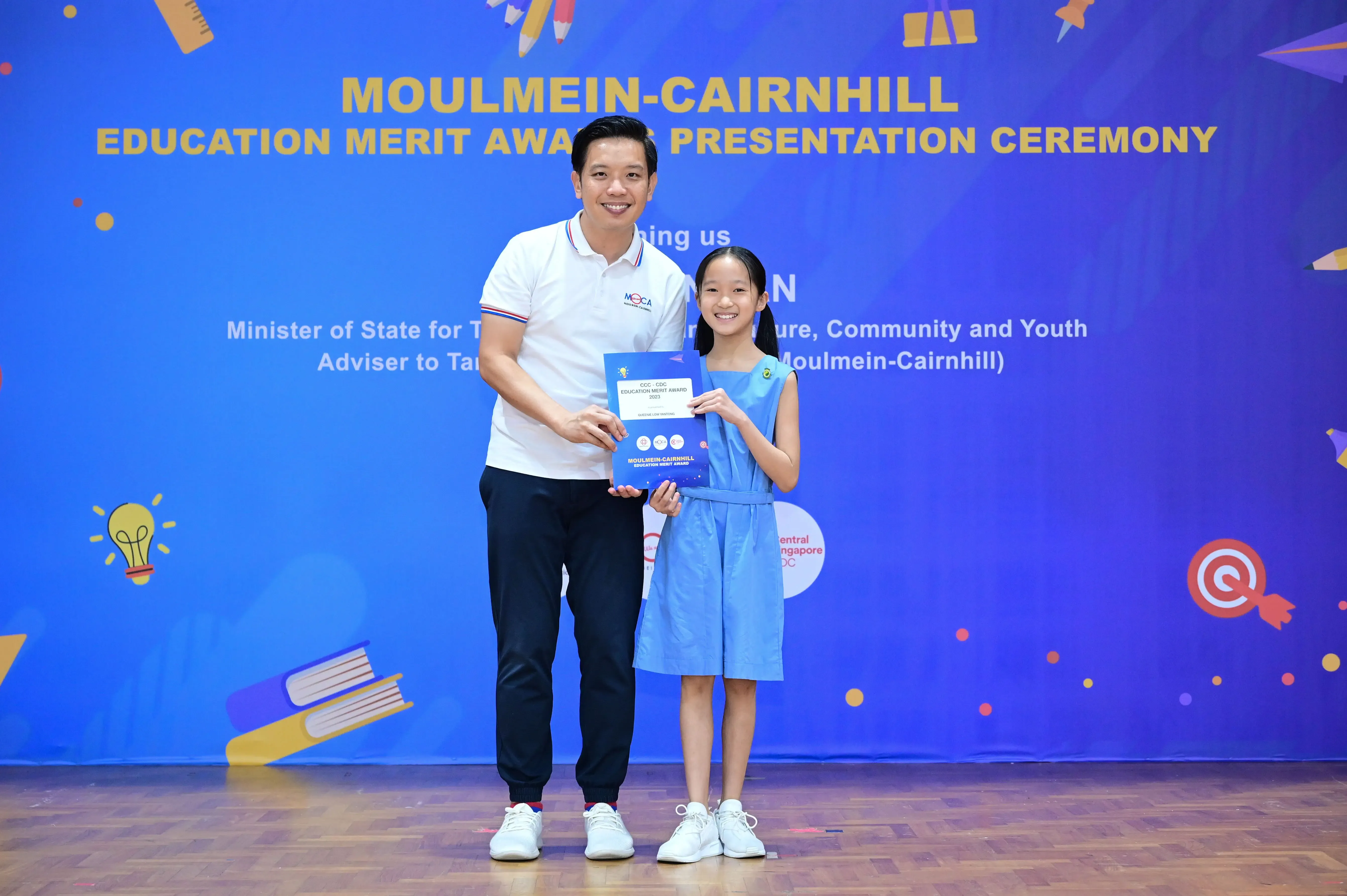 A man and a young girl hold a blue award certificate at the Moulmein-Cairnhill Education Merit Award Ceremony.