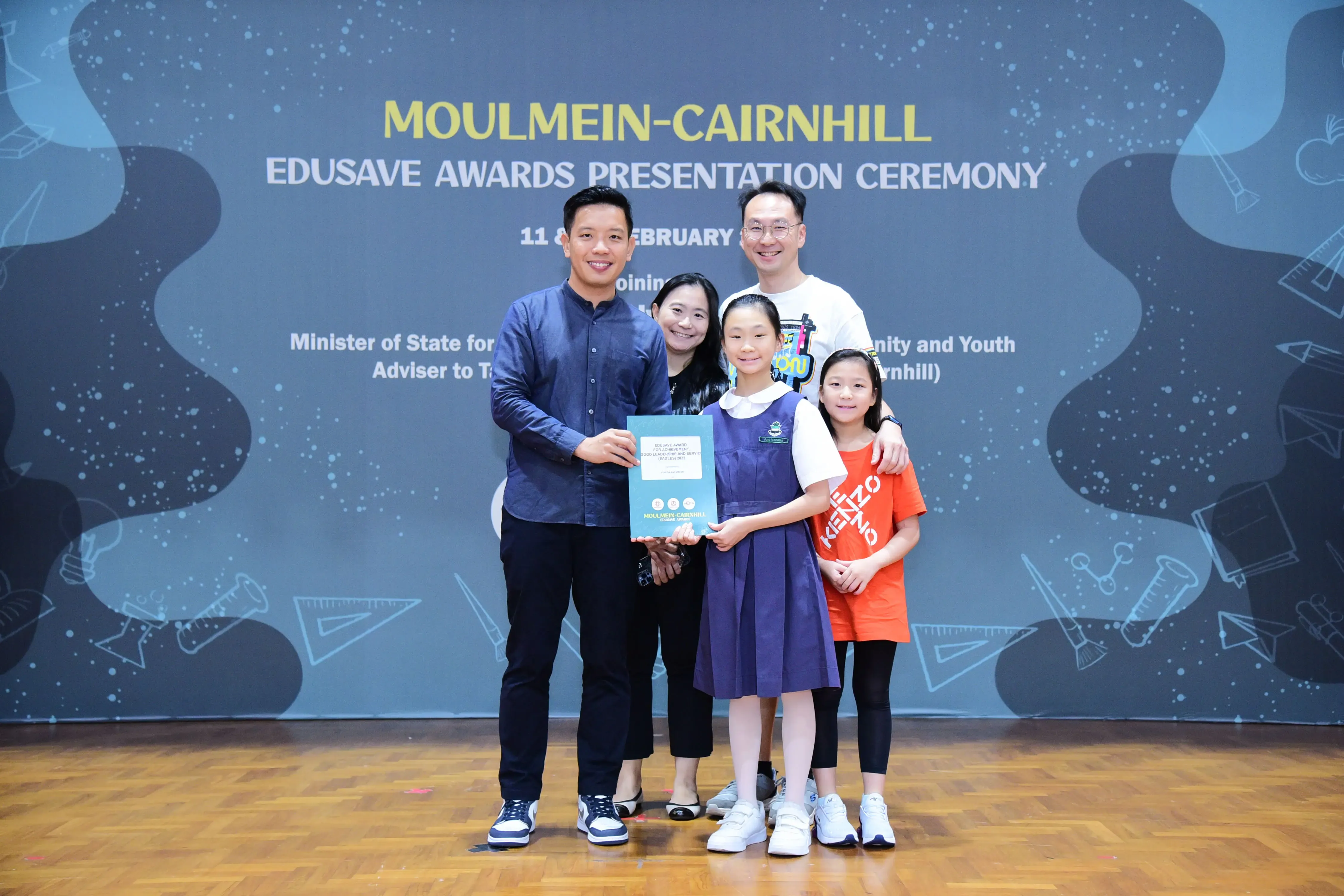 Five people pose with a certificate at the Moulmein-Cairnhill Edusave Awards Presentation Ceremony backdrop.