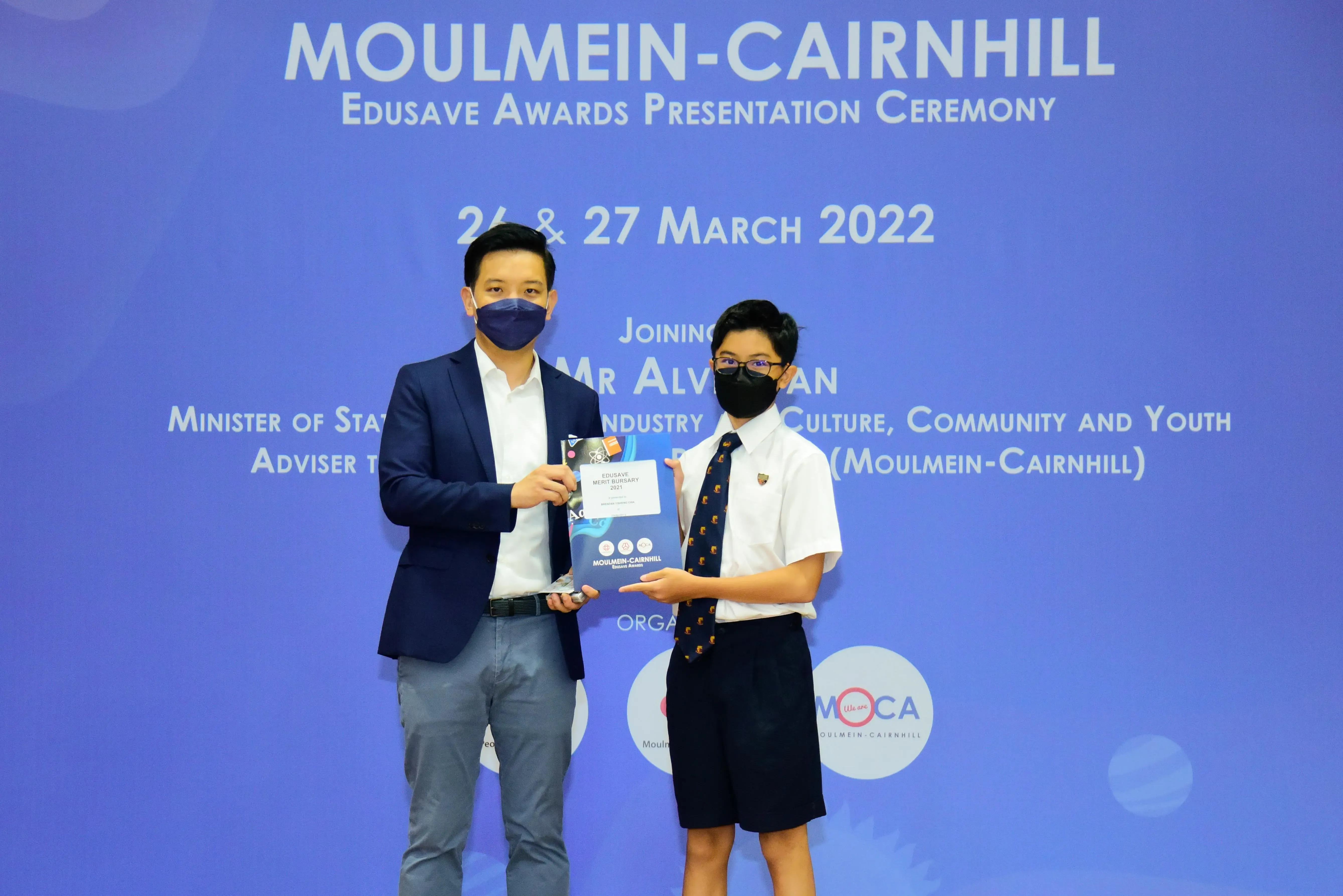 Two masked individuals hold an award folder at the Moulmein-Cairnhill Edusave Awards Presentation Ceremony, March 2022.