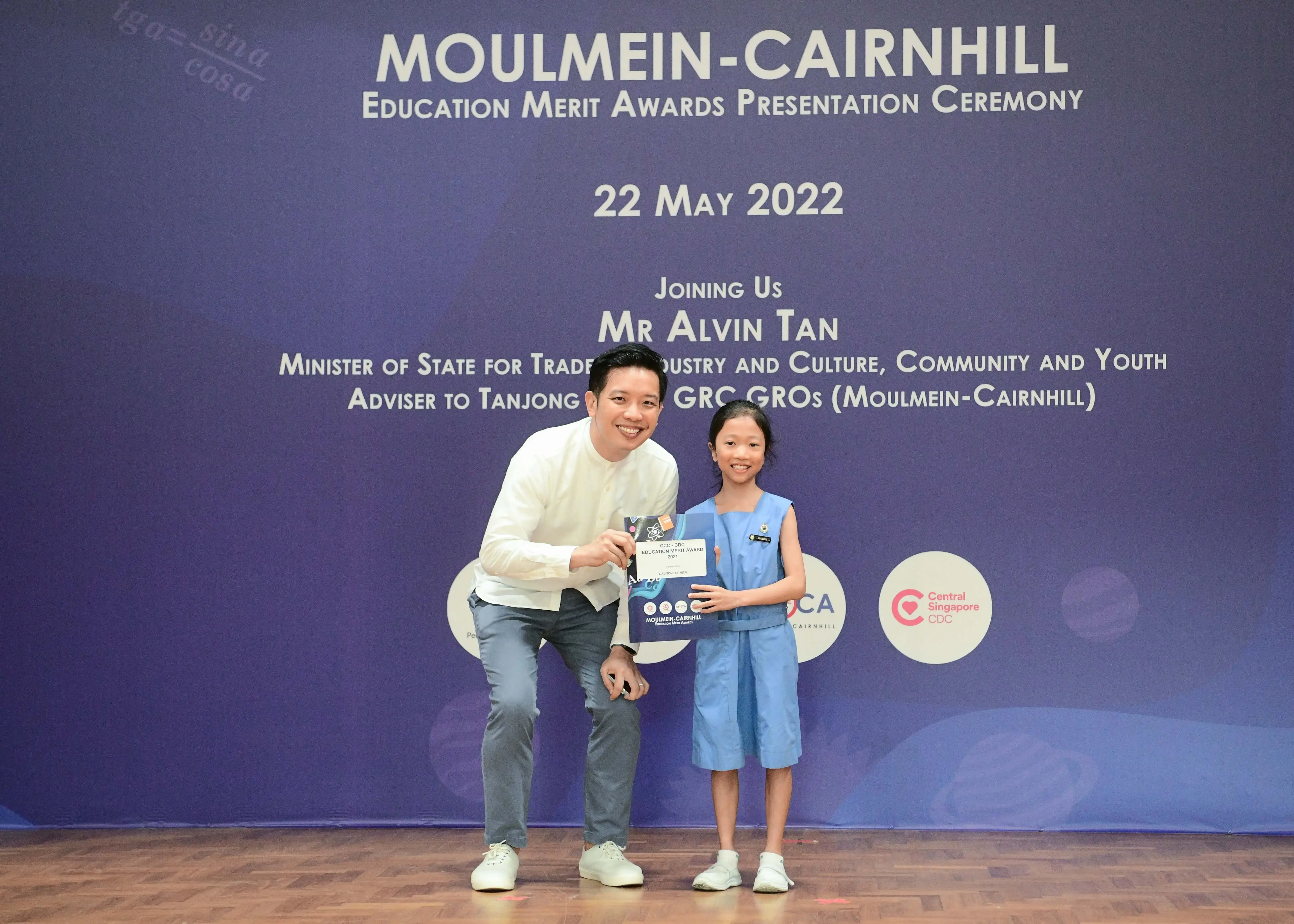 A man crouches beside a young girl holding an award certificate at the Moulmein-Cairnhill Education Merit Awards ceremony on 22 May 2022.