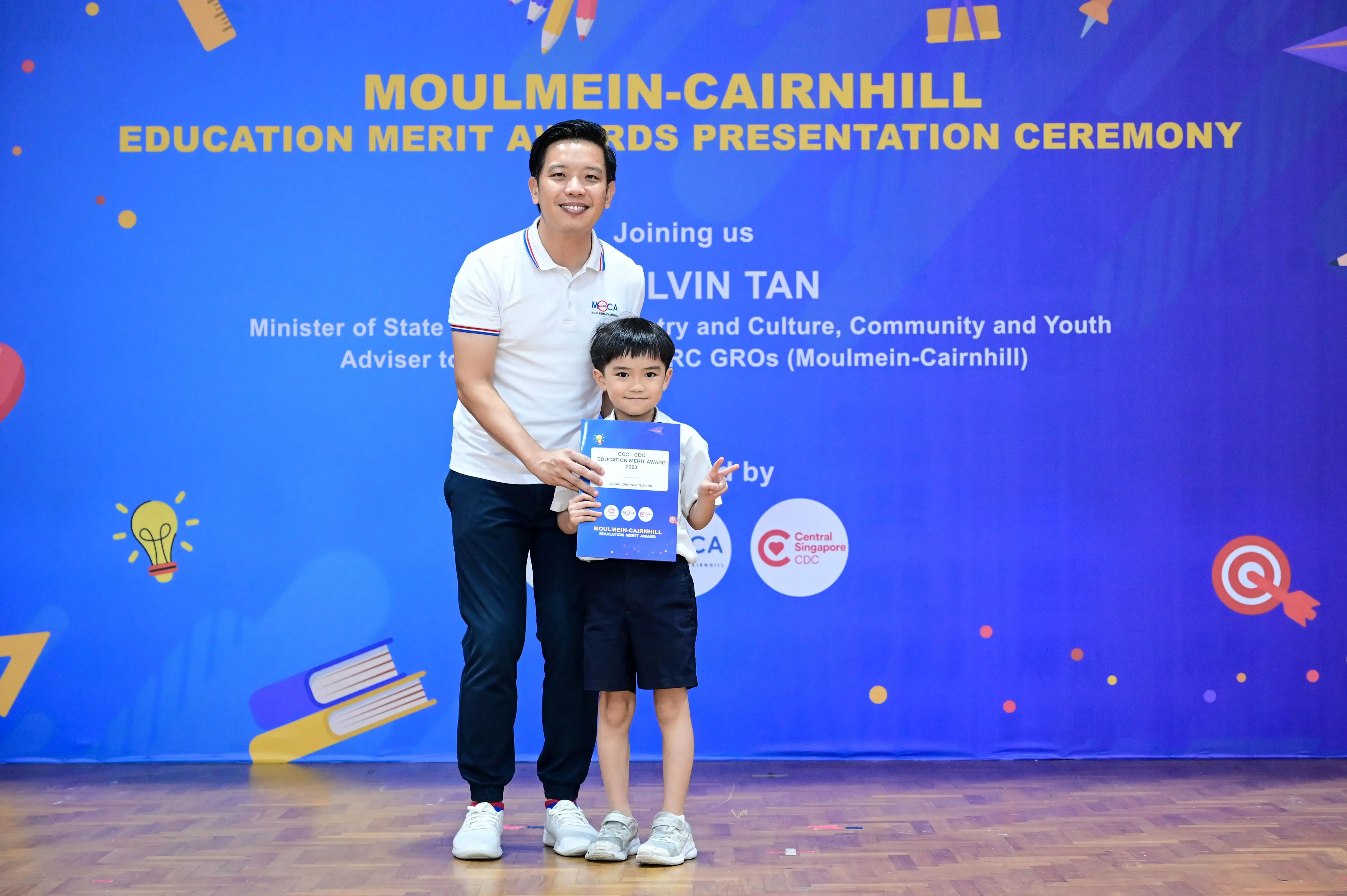 A man and young boy pose holding a certificate in front of a Moulmein-Cairnhill Education Merit Awards backdrop.