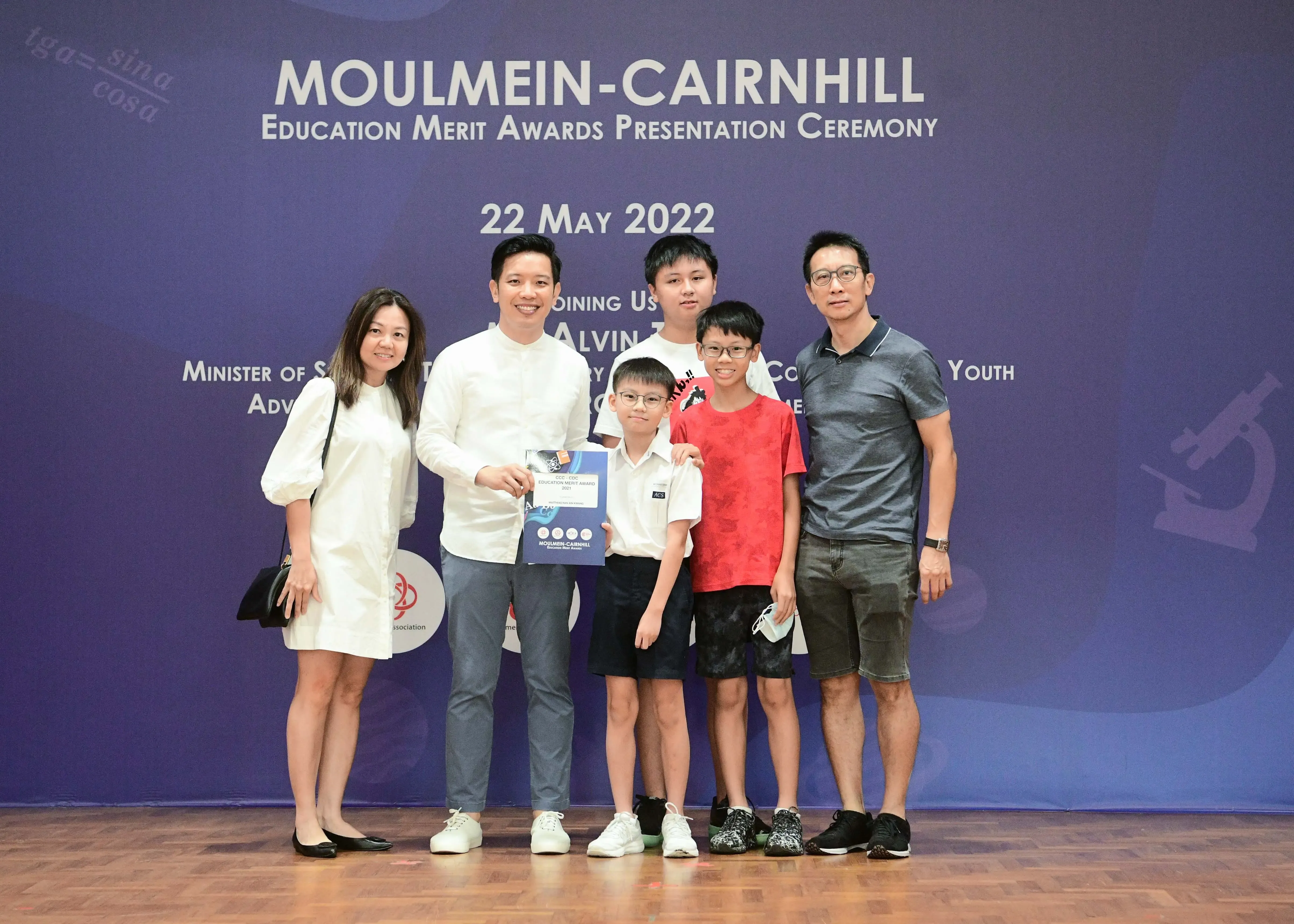 Six people pose in front of a Moulmein-Cairnhill Education Merit Awards Presentation Ceremony banner dated 22 May 2022.