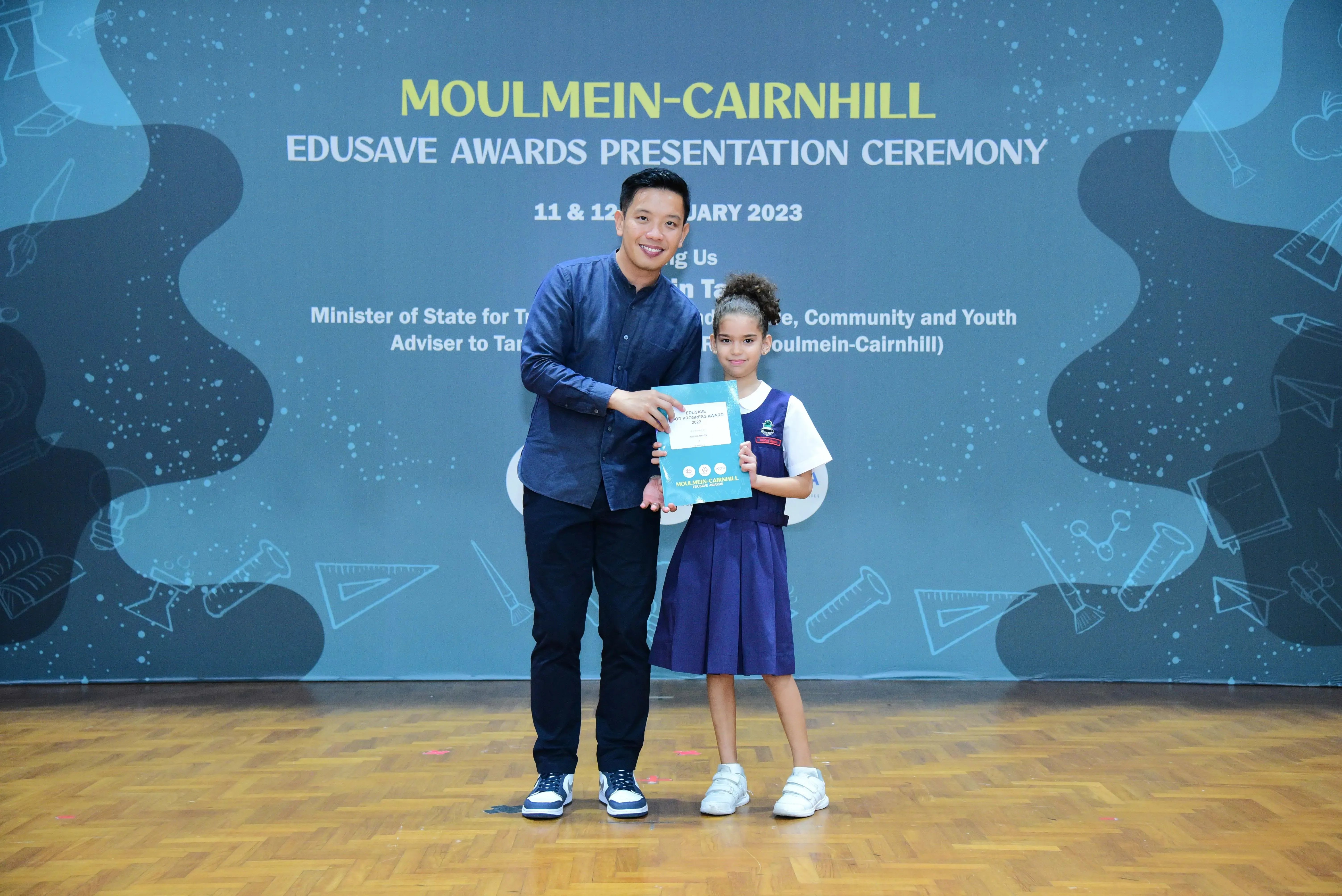 A man and a young girl in school uniform hold a certificate at the Moulmein-Cairnhill Edusave Awards Ceremony 2023.