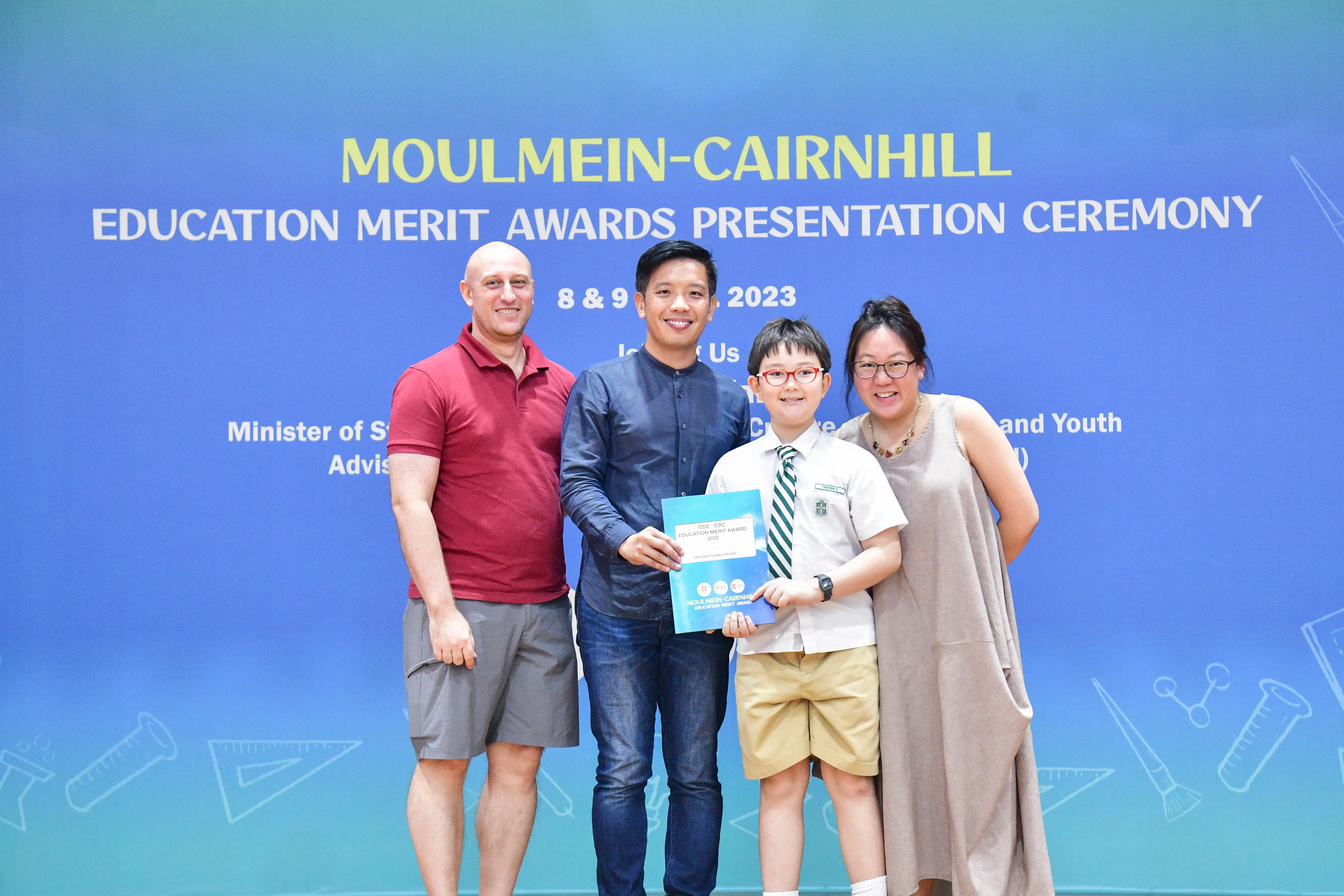Four people pose before a Moulmein-Cairnhill Education Merit Awards Presentation Ceremony banner in 2023.