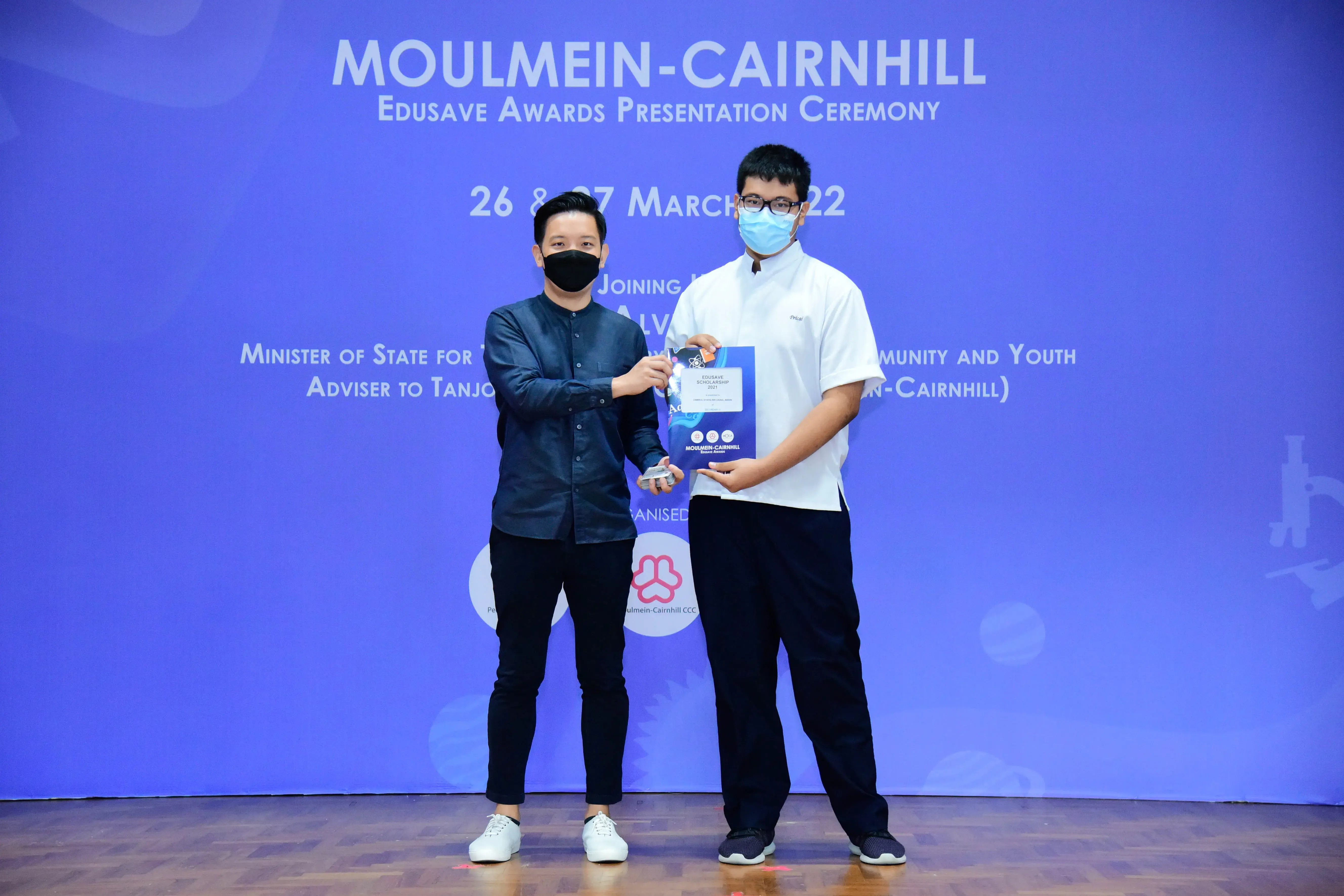 Two masked men exchange an award folder on stage at the Moulmein-Cairnhill Edusave Awards Ceremony, March 2022.