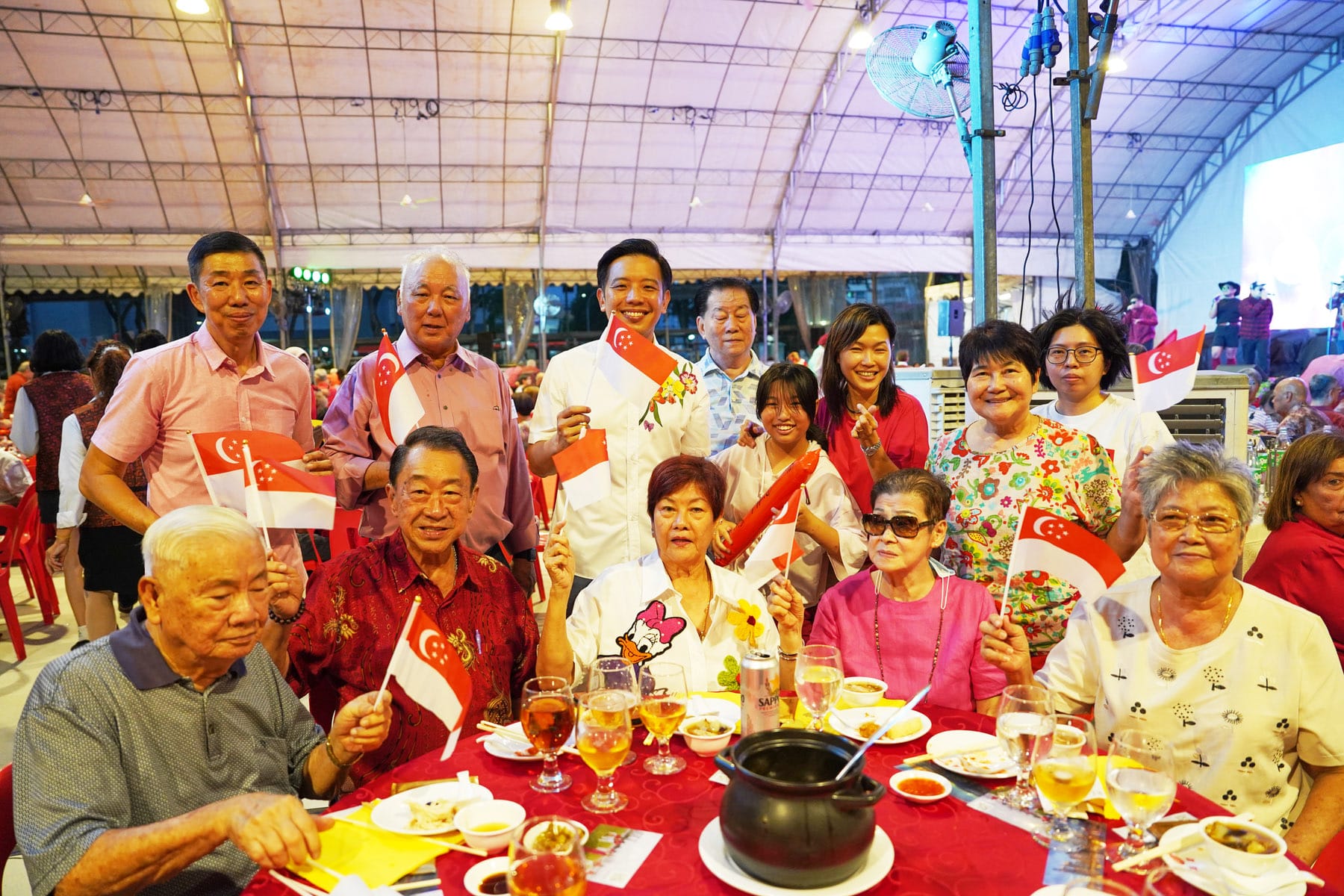 A group of smiling people wave Singapore flags at a festive dinner under a tent with food and drinks on the table.