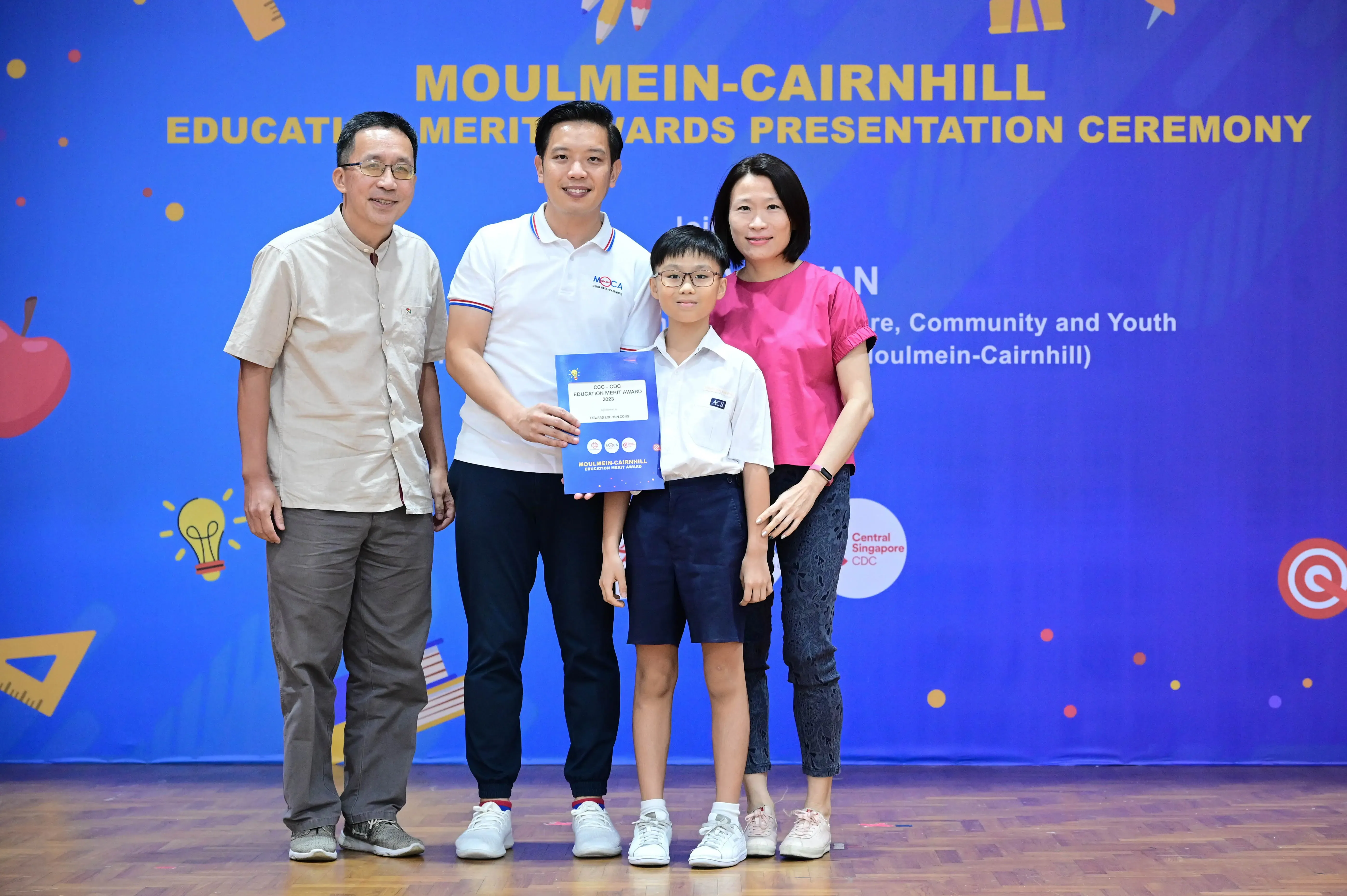 Four people pose at Moulmein-Cairnhill Education Merit Awards ceremony, a boy holding a blue award certificate.
