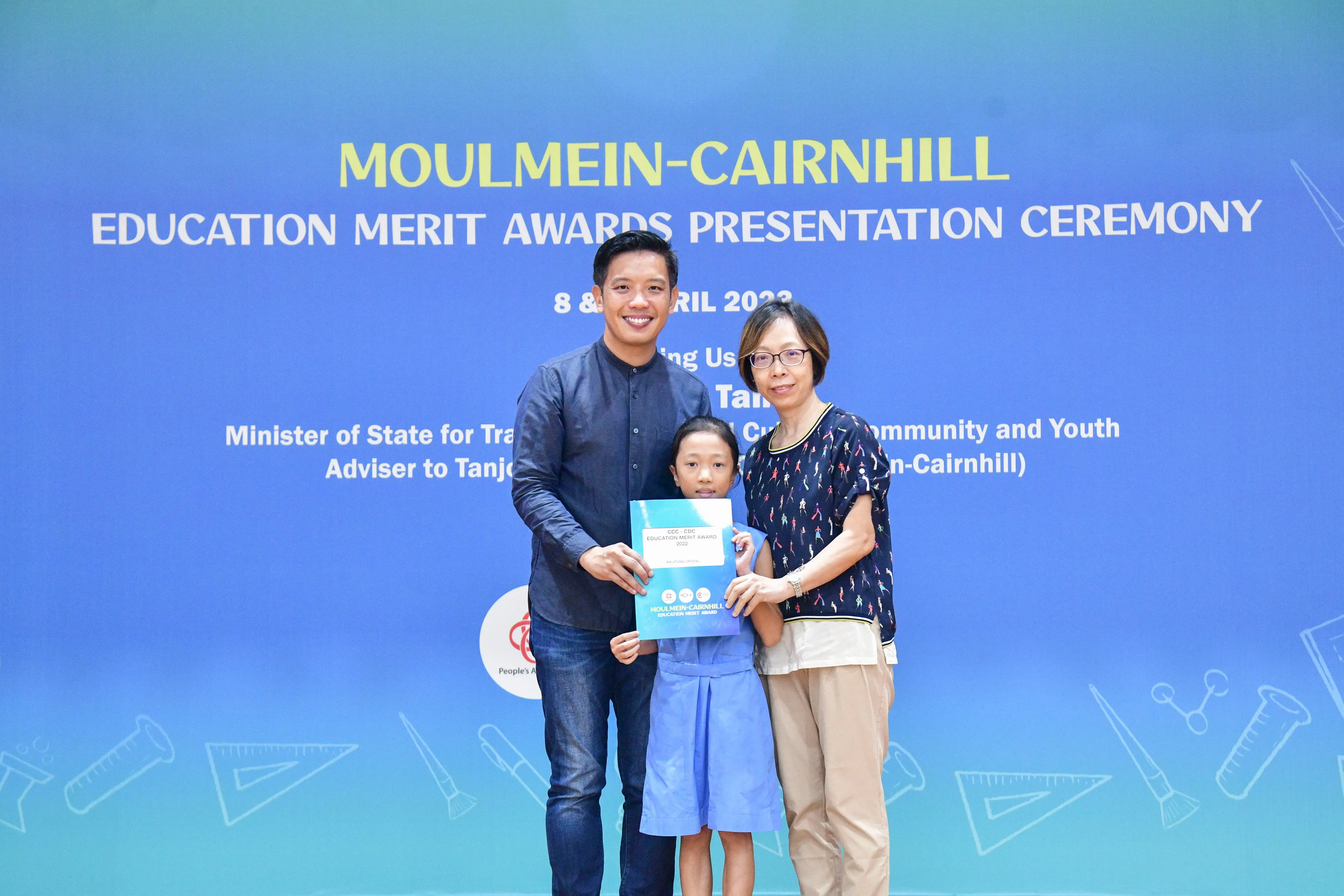 Two adults and a young girl holding an award certificate at the Moulmein-Cairnhill Education Merit Awards Ceremony.