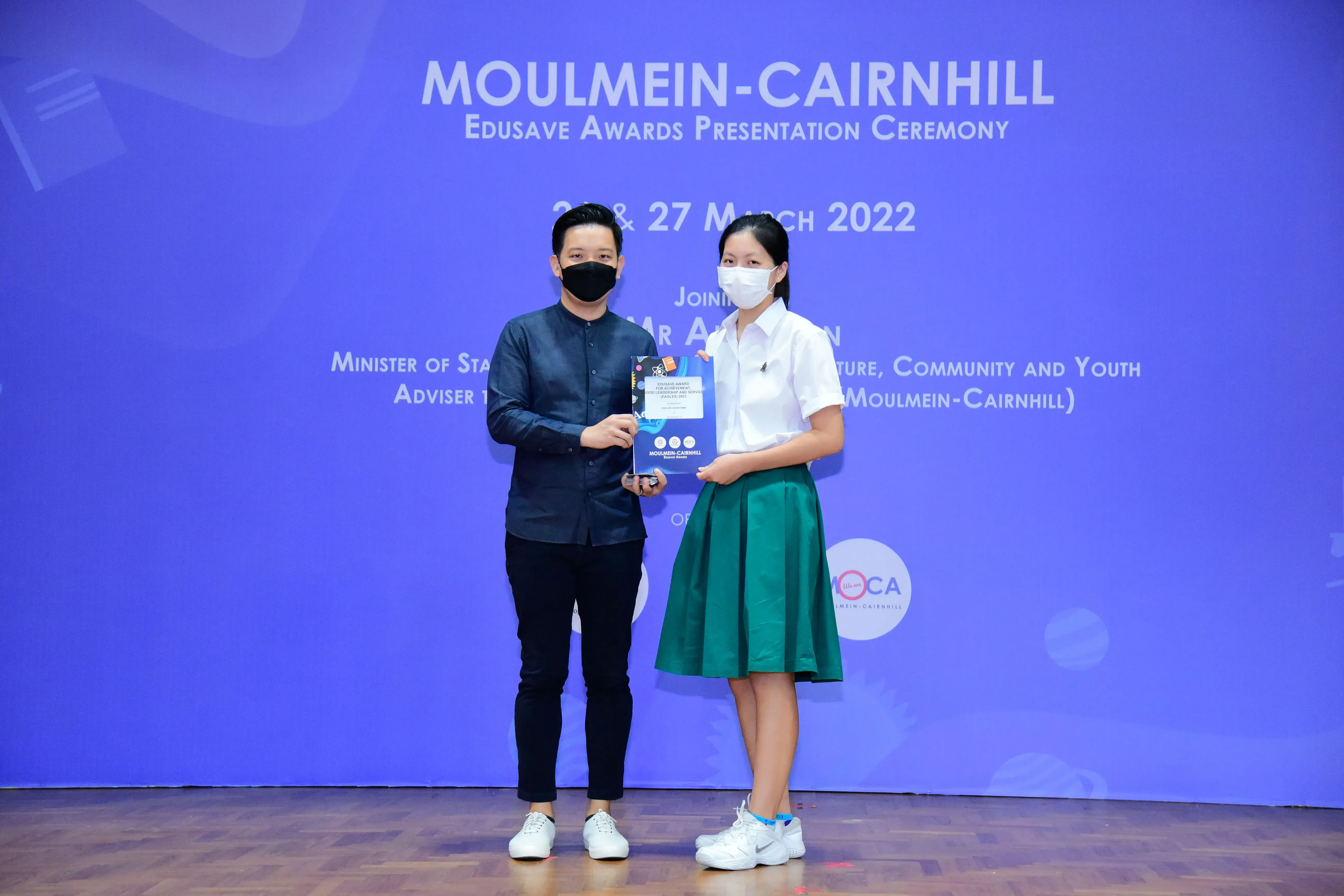 Two masked people hold an award at the Moulmein-Cairnhill Edusave Awards Presentation Ceremony, March 2022.