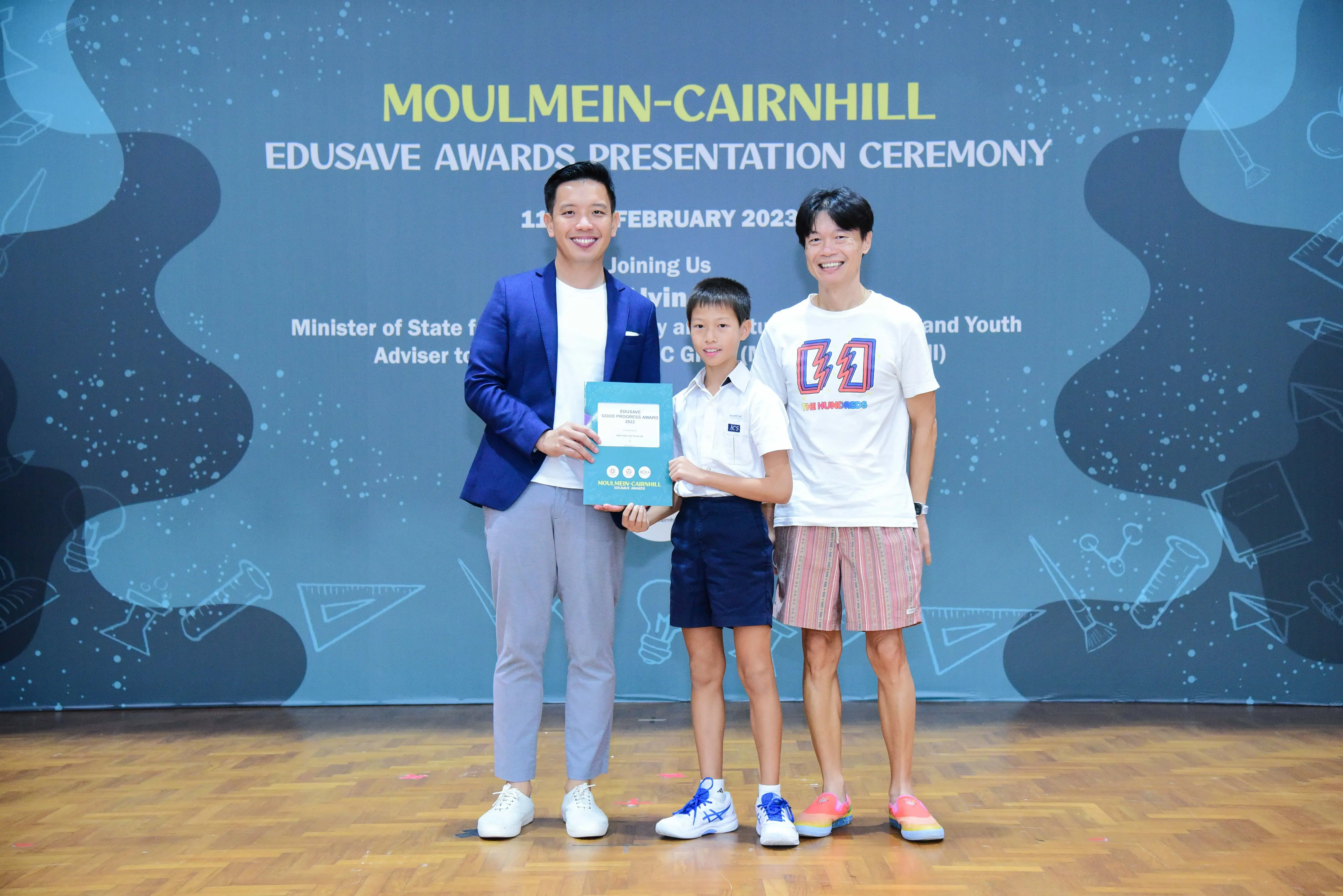 Two men and a boy hold an award booklet at the Moulmein-Cairnhill Edusave Awards Presentation Ceremony 2023.
