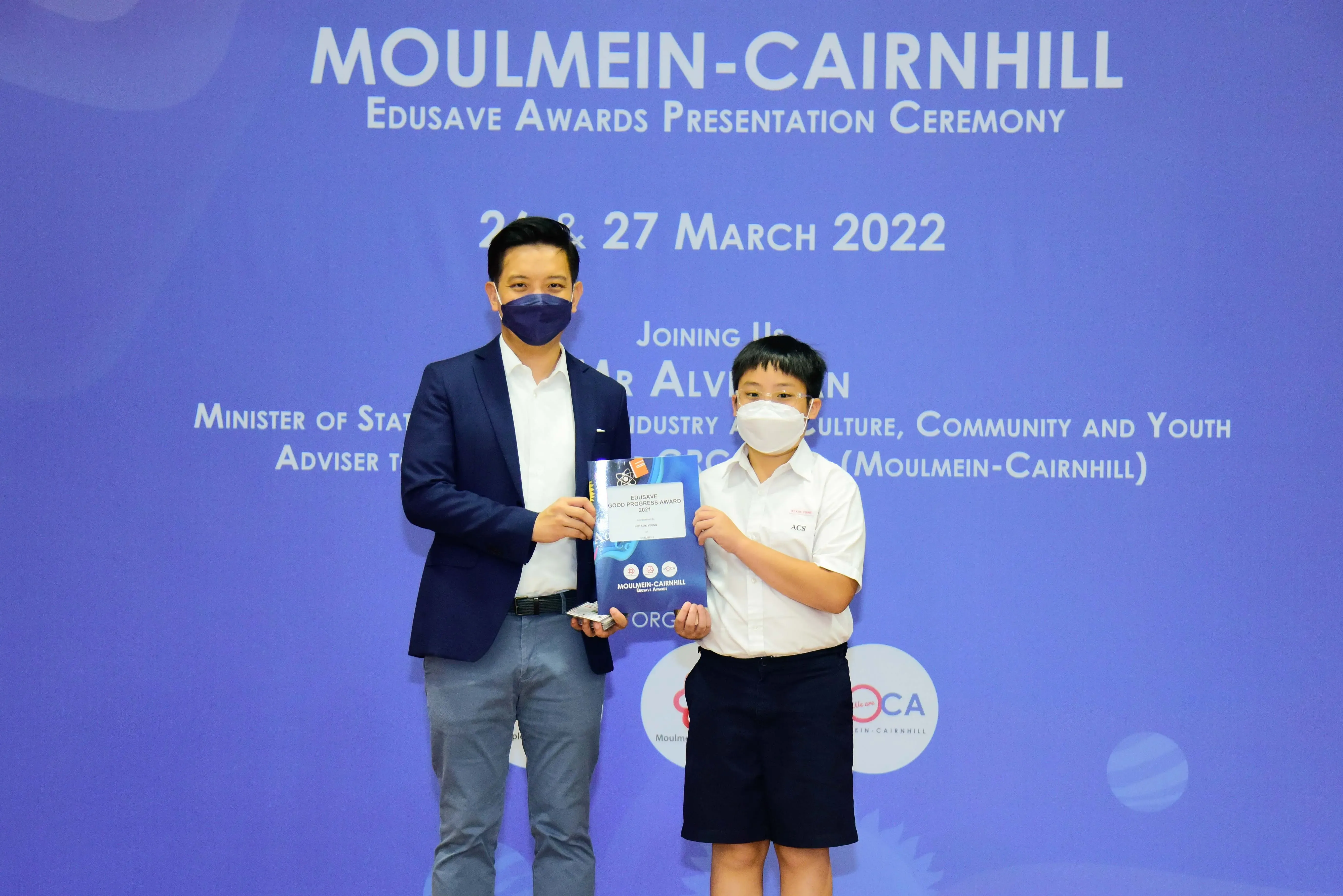 An adult and a young student in masks hold an award certificate at the Moulmein-Cairnhill Edusave Awards Ceremony 2022.