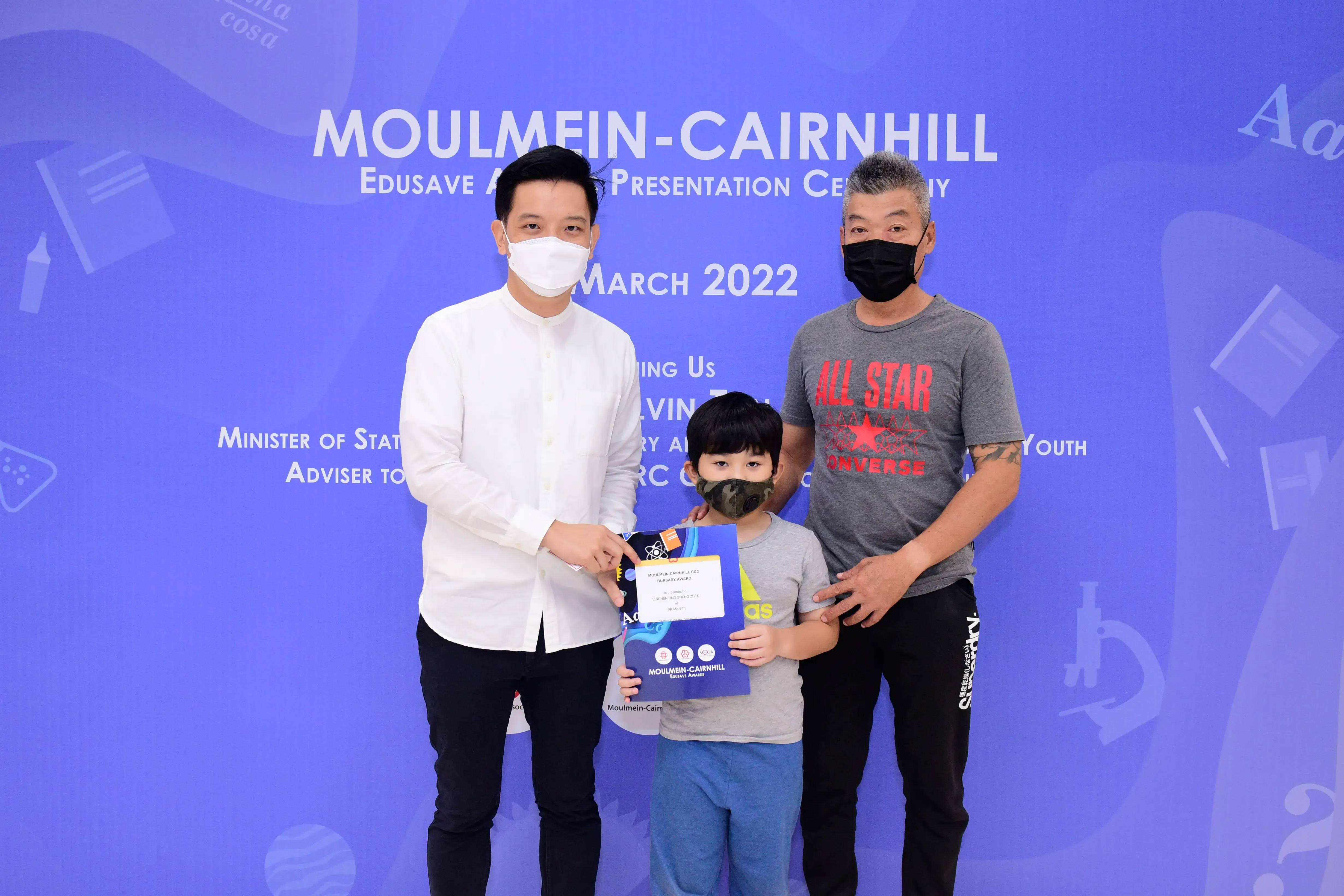 Two masked adults and a masked child holding a certificate at the Moulmein-Cairnhill Edusave Awards Ceremony, March 2022.