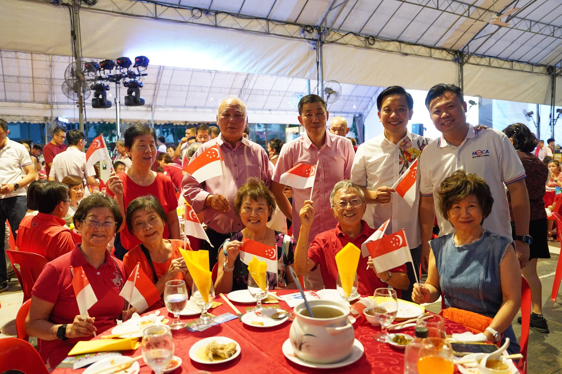 A group of smiling people holding small Singapore flags at a festive outdoor dinner under a tent.