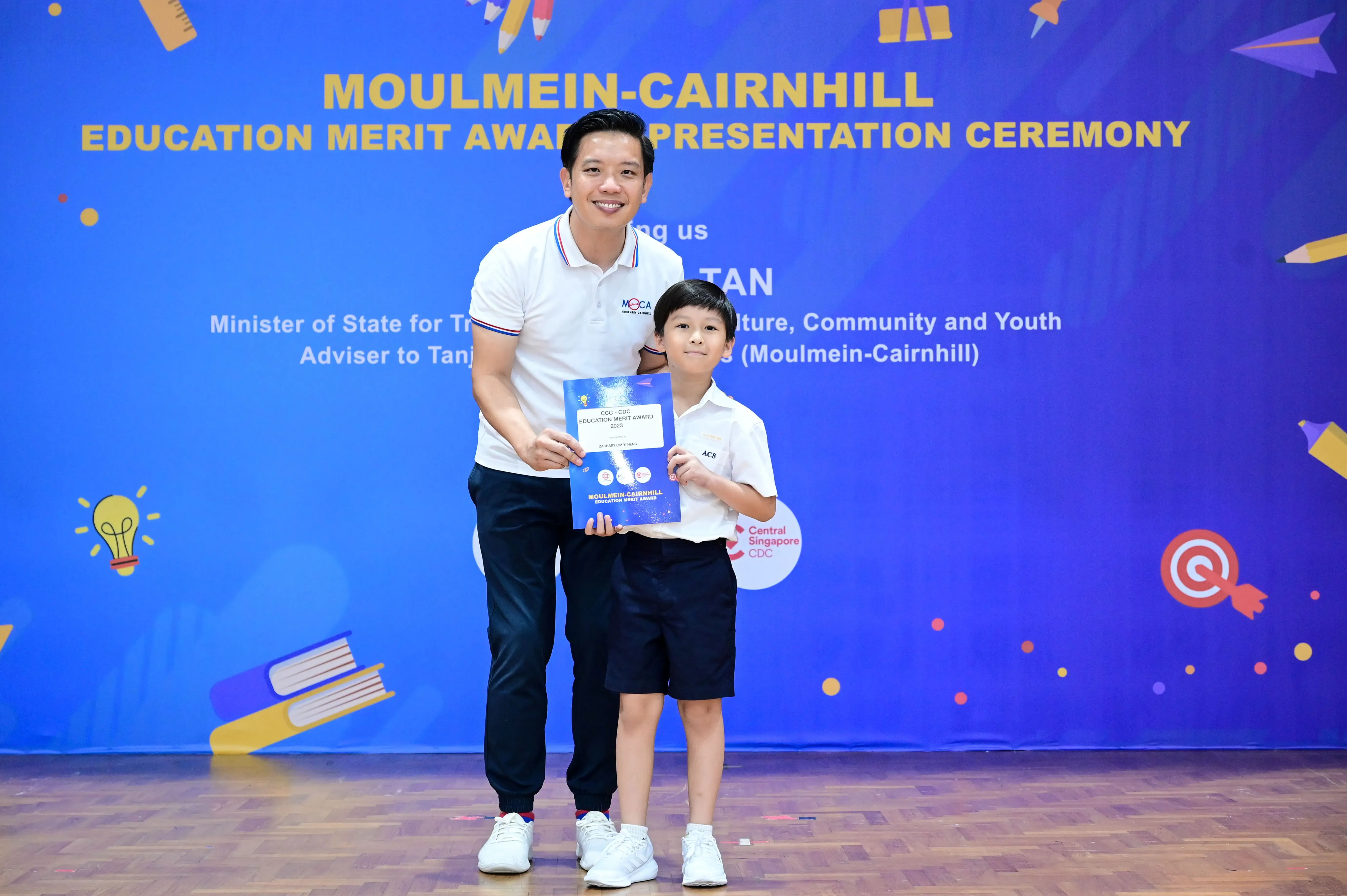 A man and young boy hold an award certificate at the Moulmein-Cairnhill Education Merit Award Presentation Ceremony.
