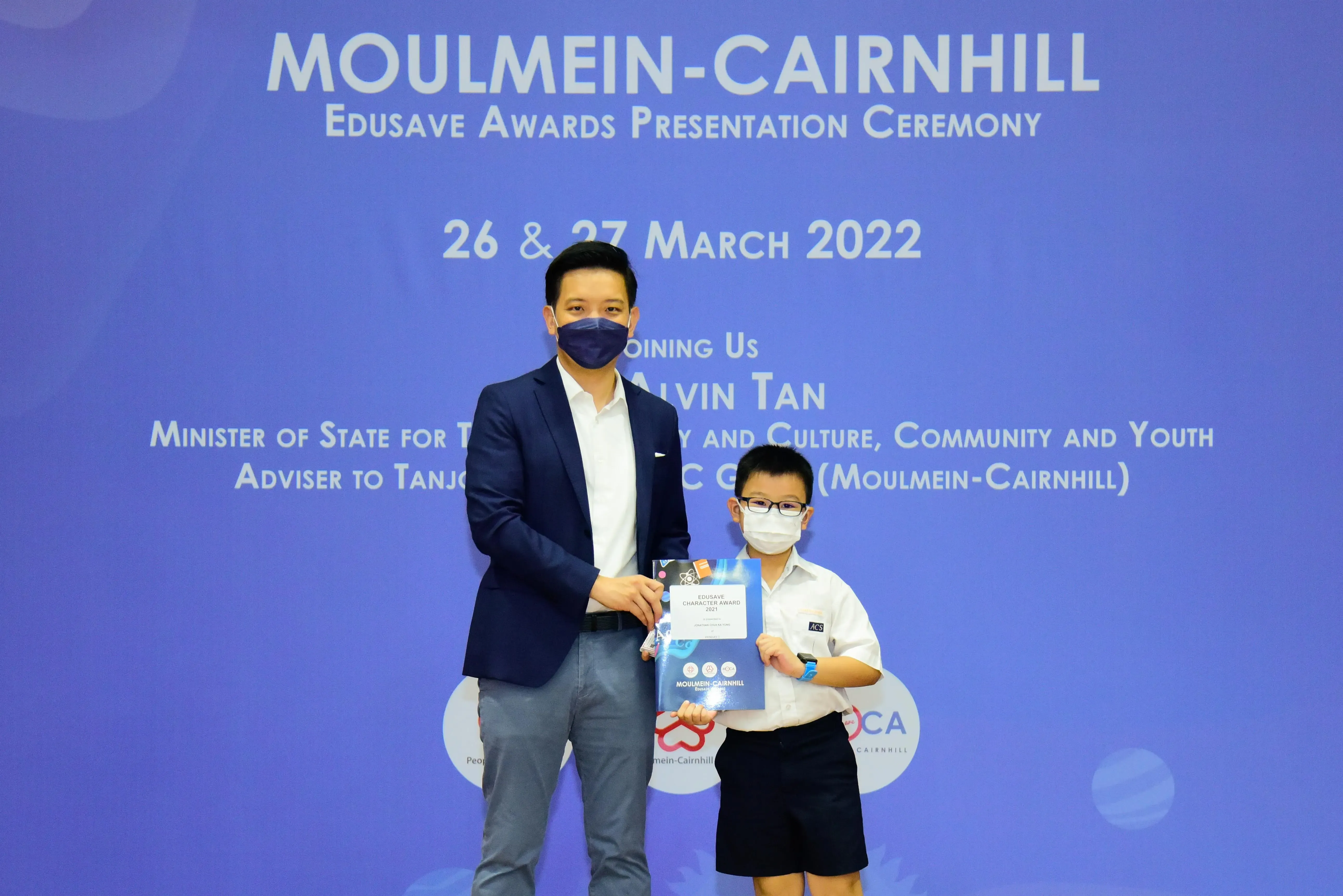 An adult and a young student in uniform hold a certificate at the Moulmein-Cairnhill Edusave Awards Ceremony, March 2022.