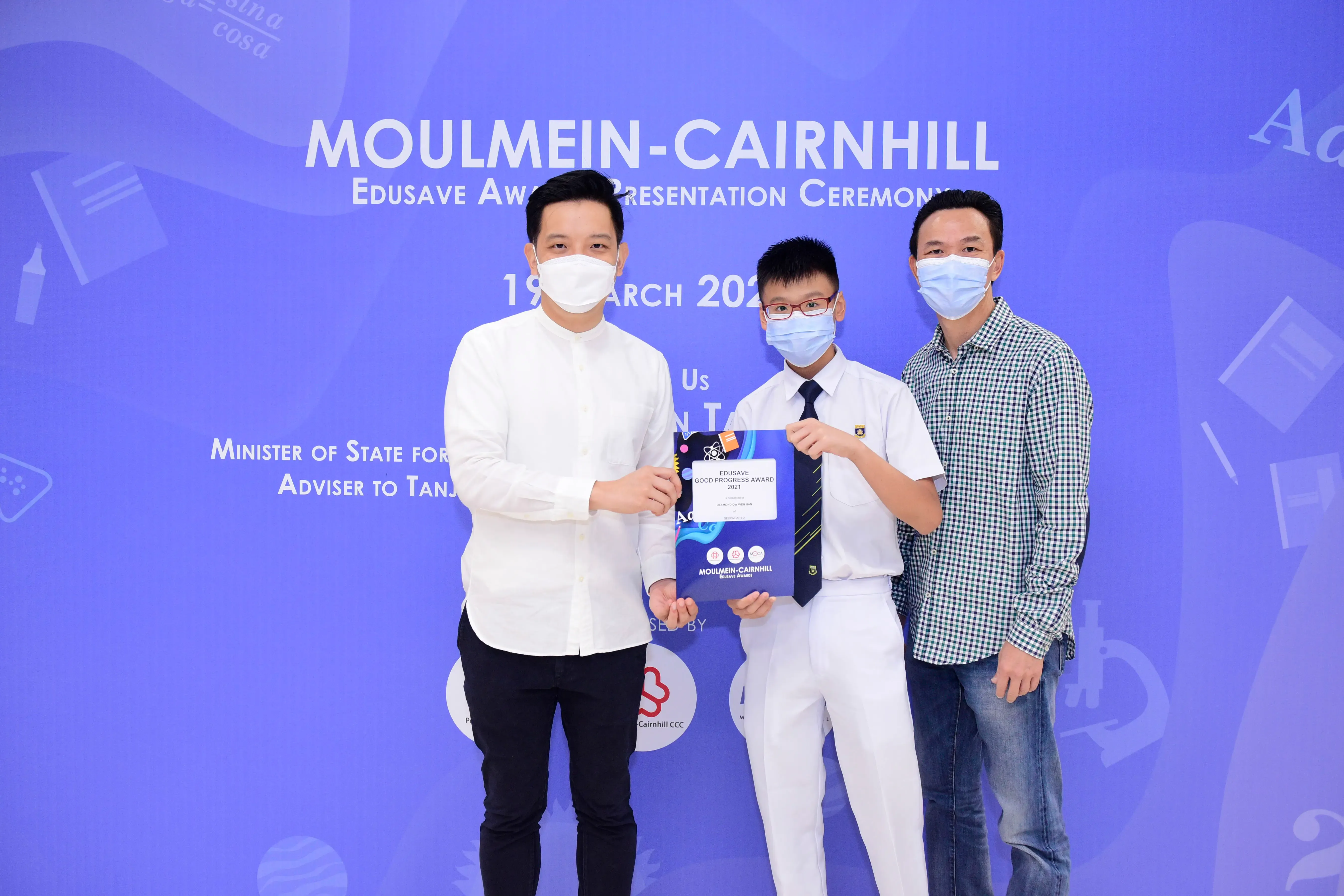Three masked people pose at Moulmein-Cairnhill Edusave Award Ceremony, with a student holding an award certificate.