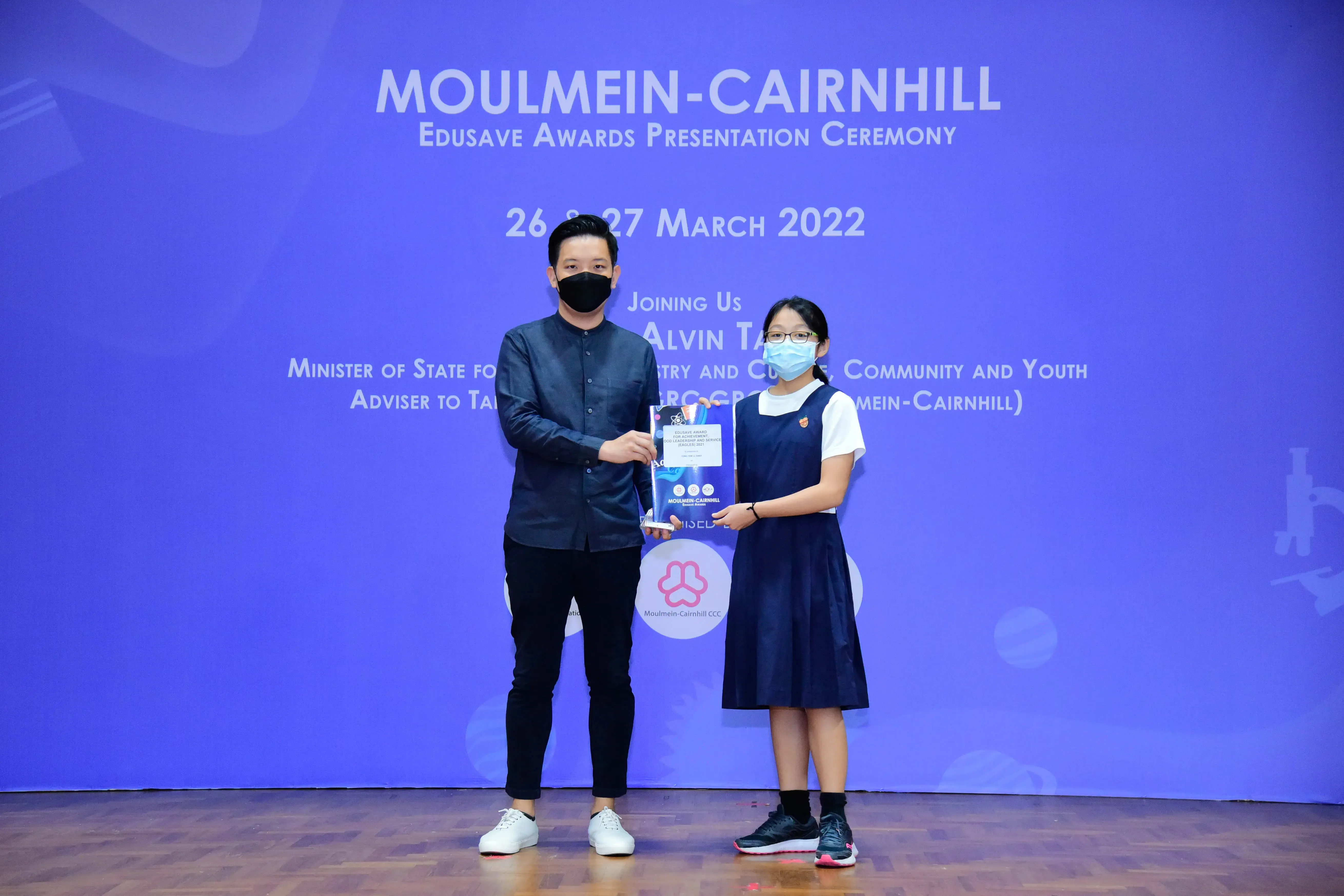 Two masked people hold a trophy on stage at the Moulmein-Cairnhill Edusave Awards Ceremony, March 2022.