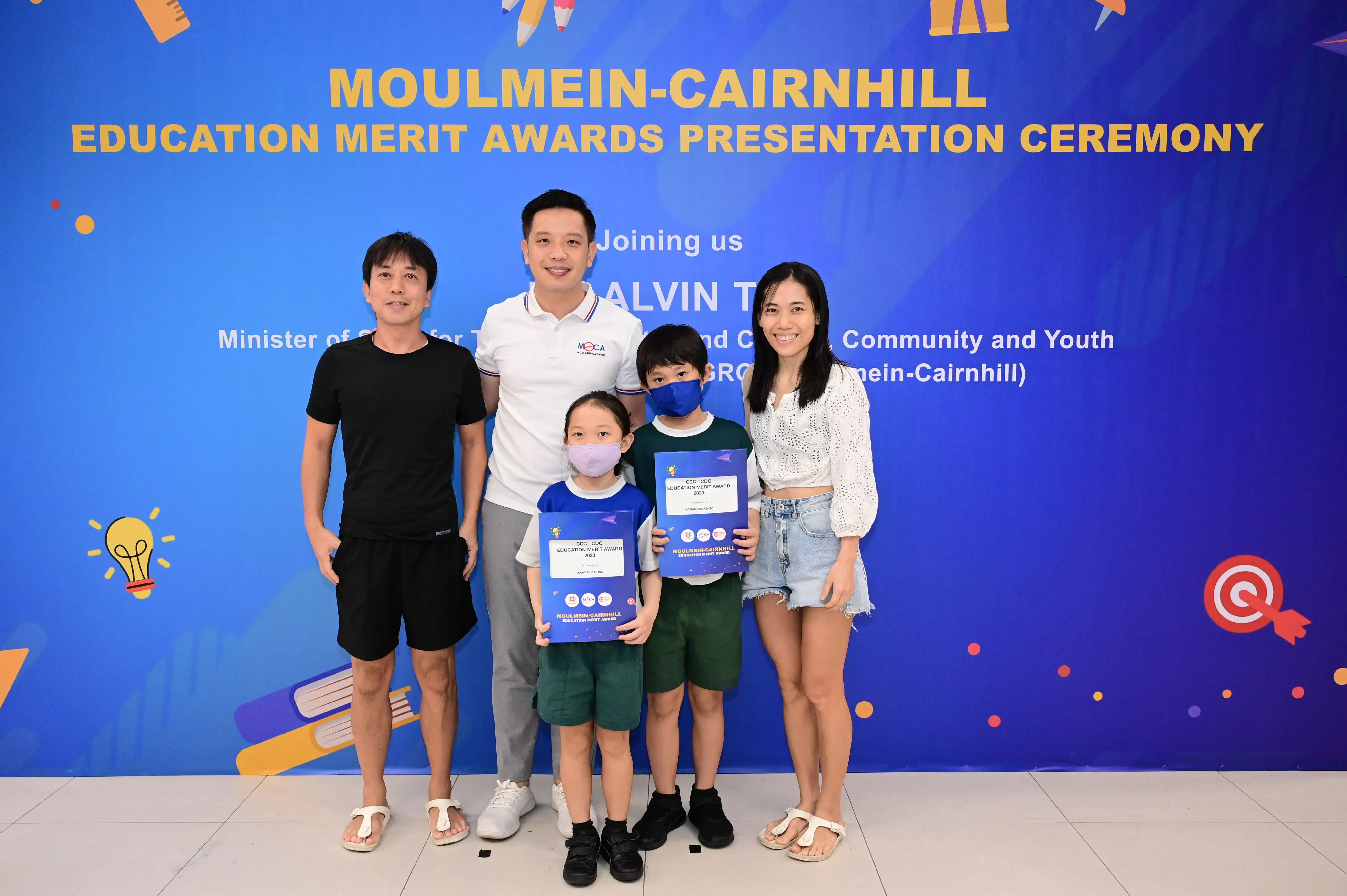 Two adults and two children holding award certificates pose before a Moulmein-Cairnhill Education Merit Awards banner.