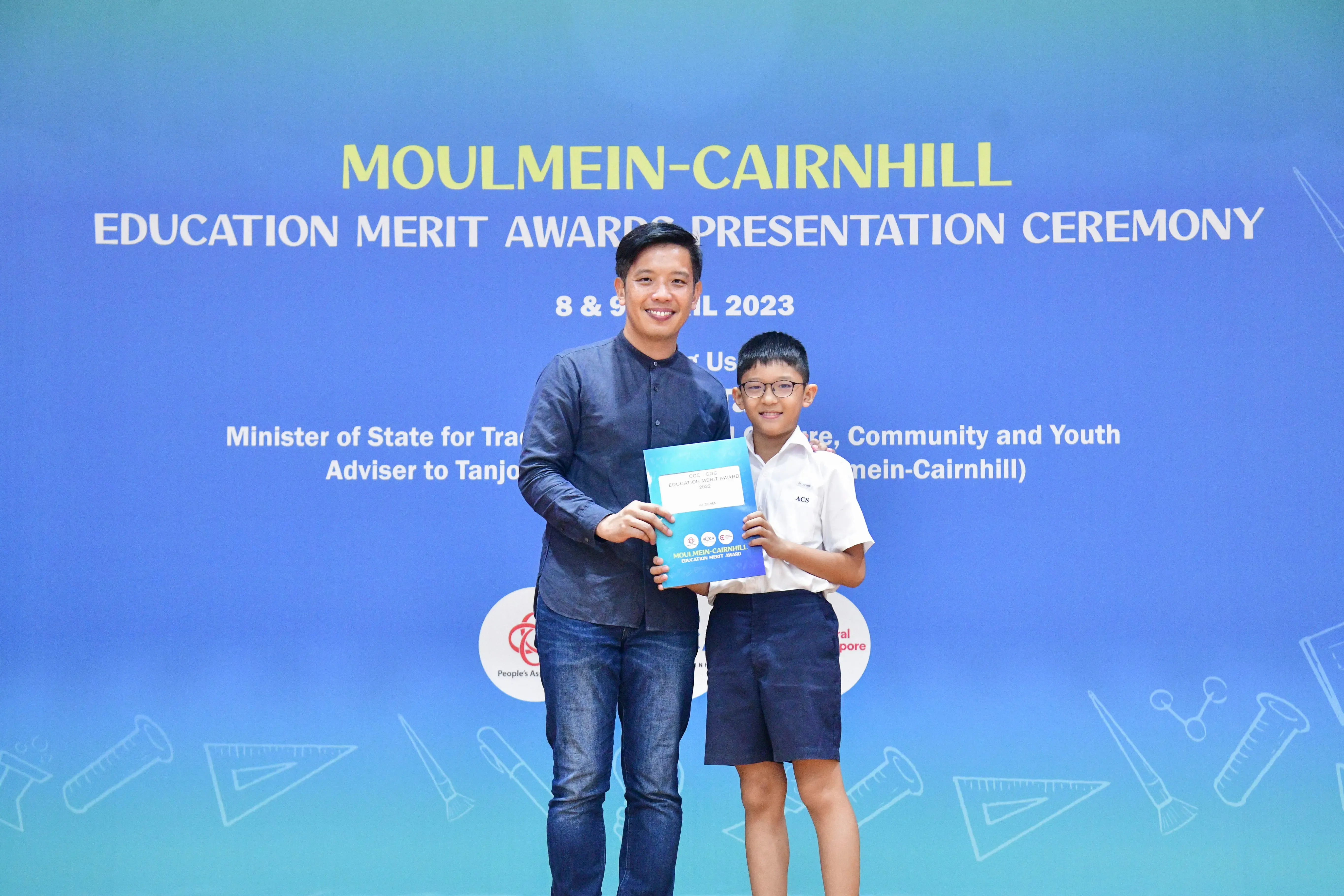 An adult man and a young boy in school uniform hold a certificate at the Moulmein-Cairnhill Education Merit Awards 2023.