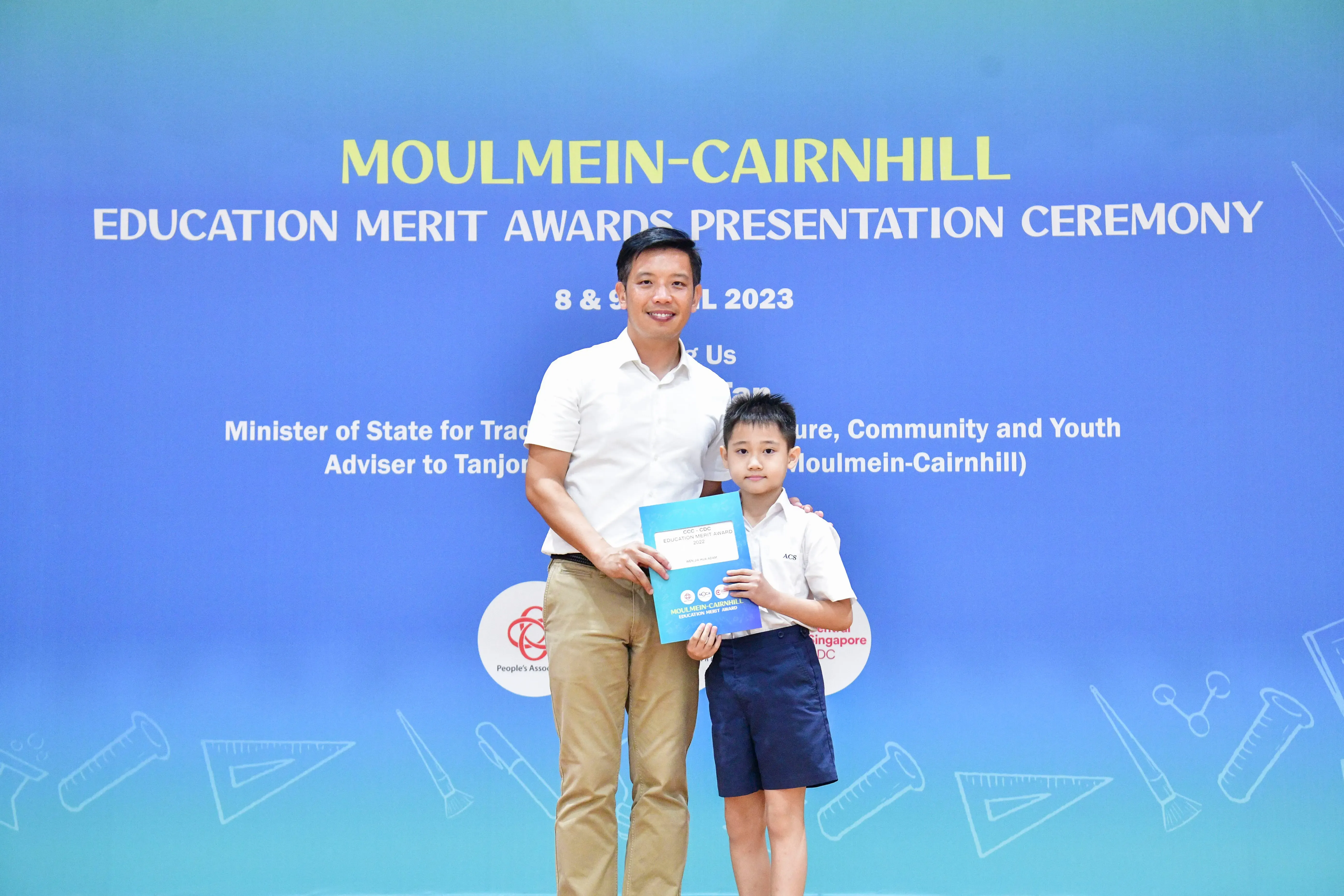 A man and a young boy hold an award certificate in front of a Moulmein-Cairnhill Education Merit Awards banner.