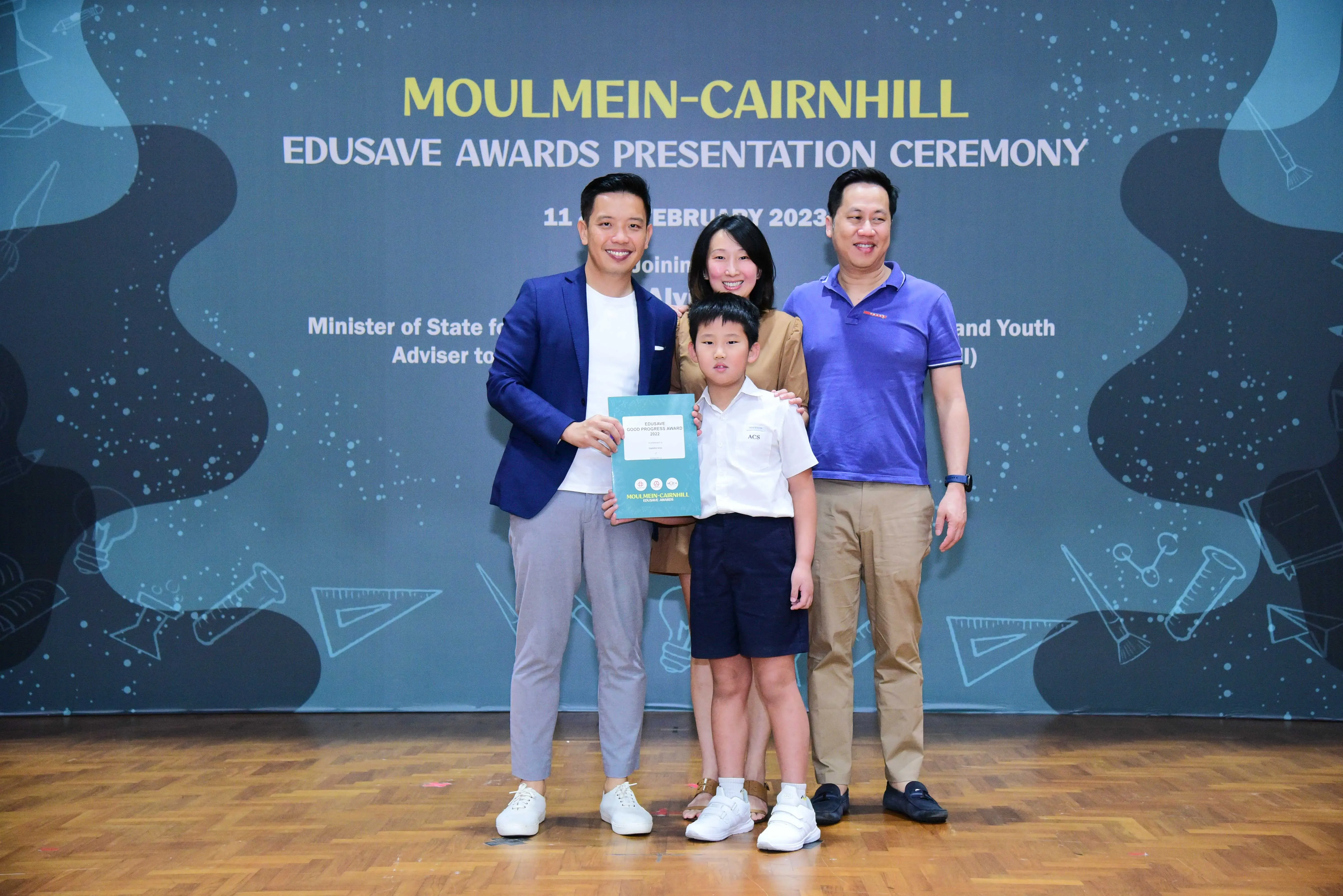 Four people pose at the Moulmein-Cairnhill Edusave Awards Presentation Ceremony, with a boy holding a teal certificate.