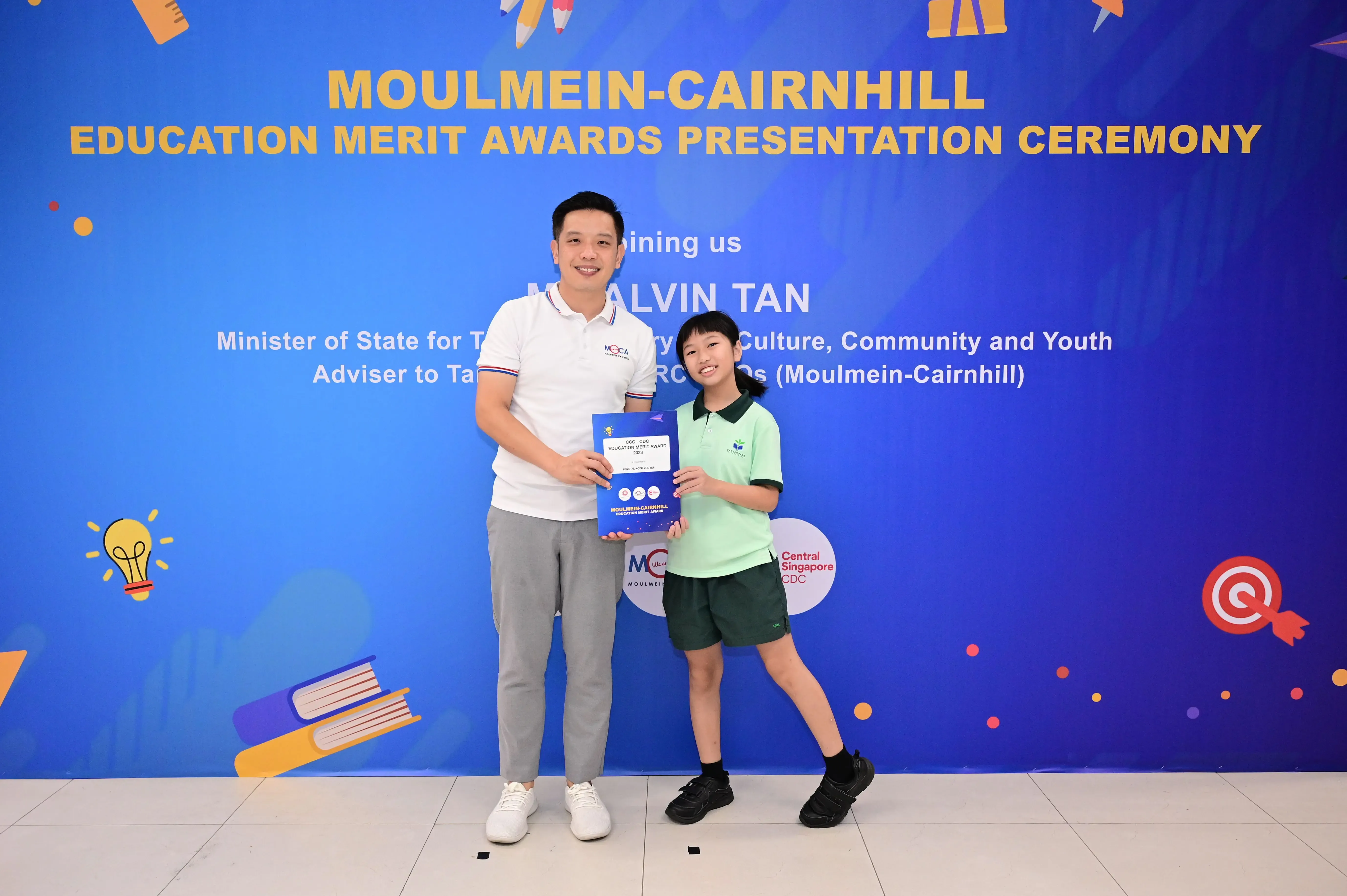 A man and a young girl hold an award certificate in front of a Moulmein-Cairnhill Education Merit Awards banner.
